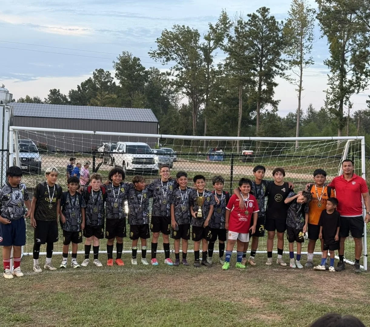 A youth soccer team wearing black and grey uniforms with medals around their necks, standing in a line on a soccer field with a goal behind them, holding a trophy, with a coach on the right, smiling. There are trees, parked cars, and a building in th