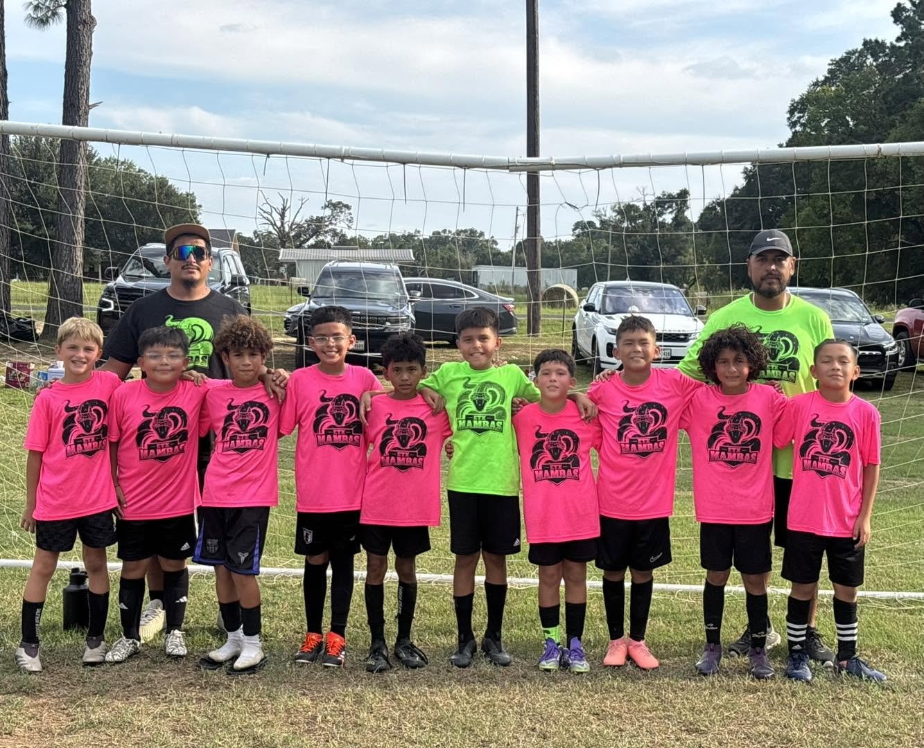 Children and coaches on a soccer field team photo, kids in pink jerseys with black shorts, coaches in neon green and black shirts, standing in front of a soccer net with parked cars and trees in the background.