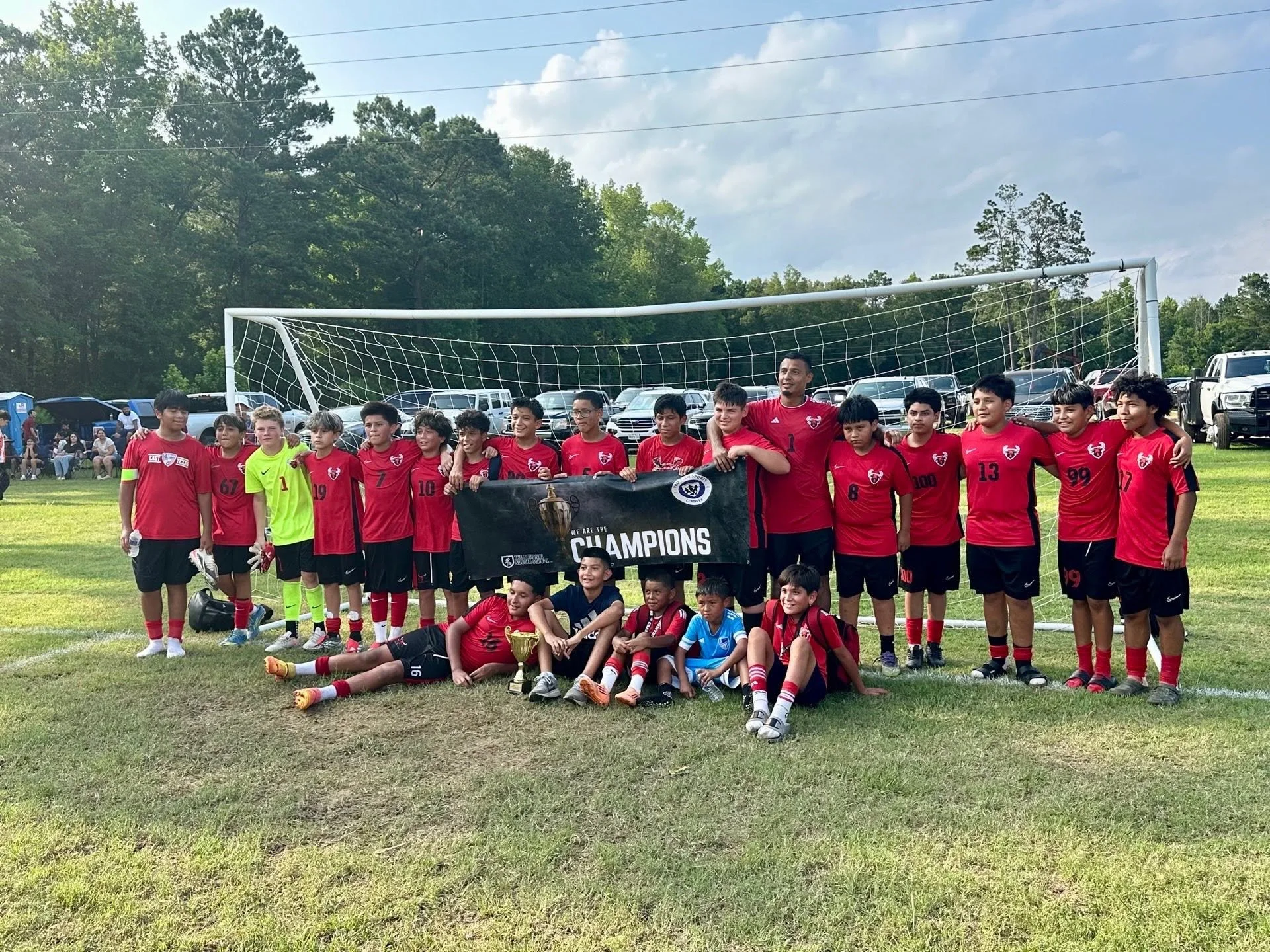 A youth soccer team in red jerseys and black shorts, with some players wearing goalkeeper uniforms, stands and sits on a soccer field, holding a large black banner that reads 'We Are the Champions' with a trophy image. They are posing in front of a soccer goal, with other people in the background, and parked cars behind them.