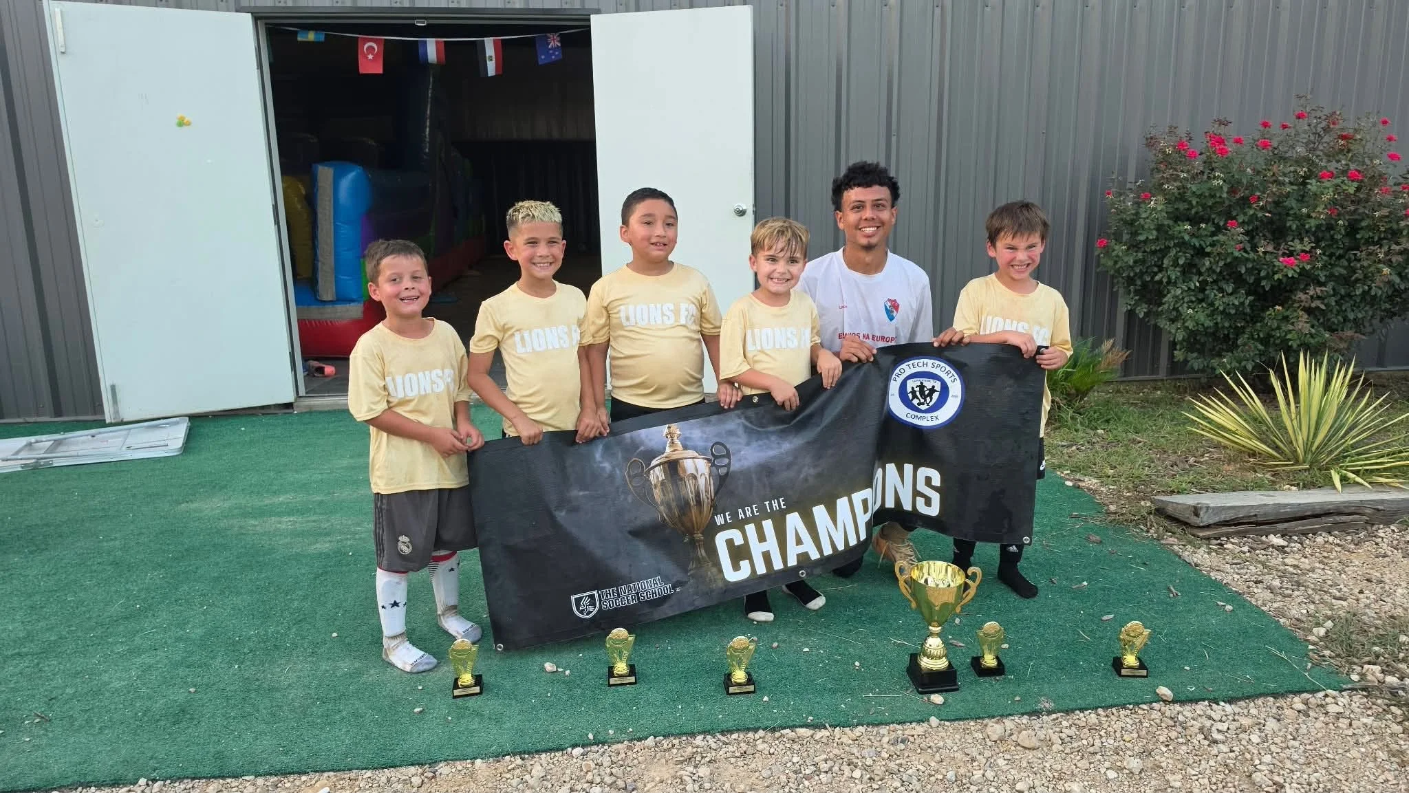 A group of young boys and a teenage man holding a banner that reads 'We Are the Champions' at a soccer awards ceremony, with trophies placed on the ground in front of them, outside near a shed with flowers and flags.