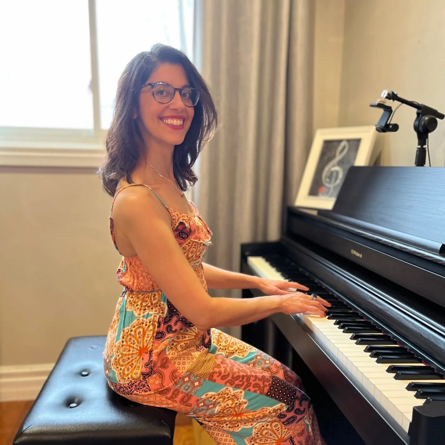 Nefeli sitting at her piano, smiling, wearing a colourful floral jumpsuit.