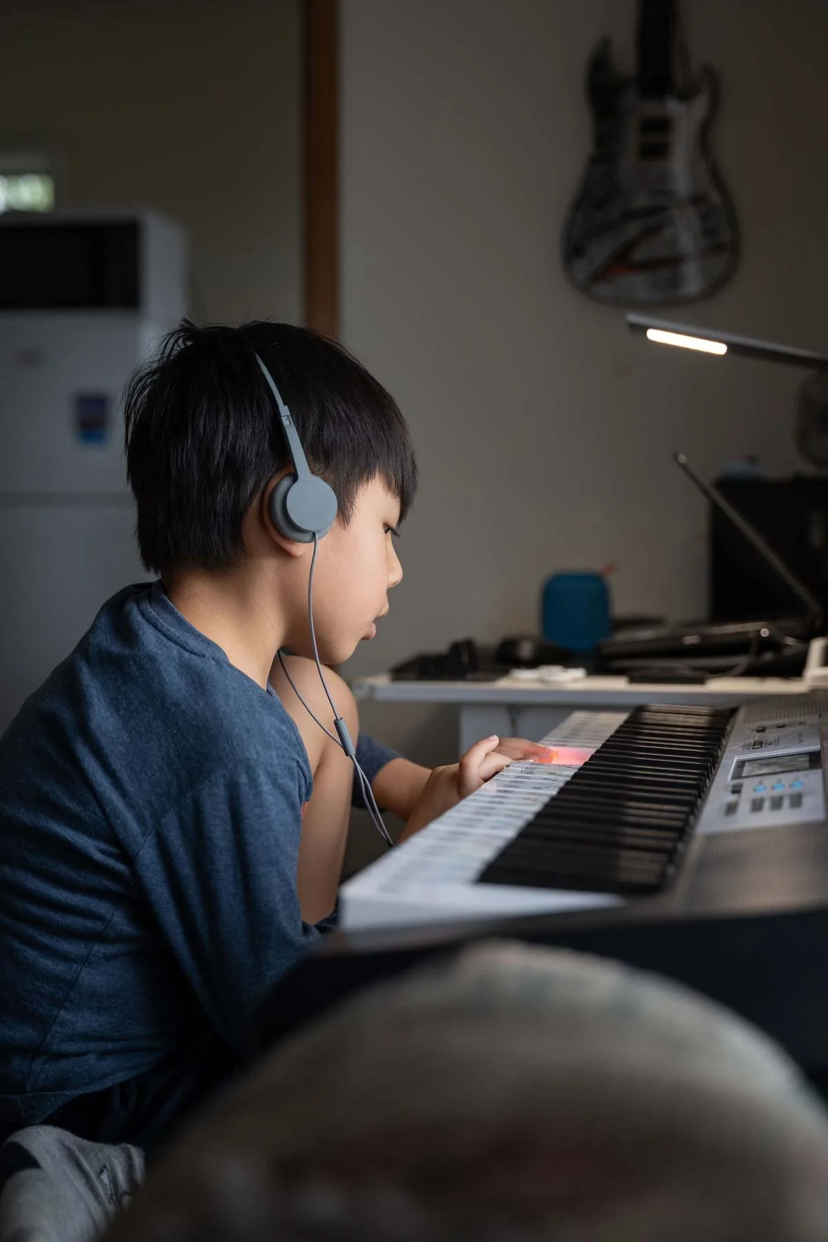 Side view of a young Asian boy wearing headphones, sitting with his knees up to his chin while playing the keyboard.