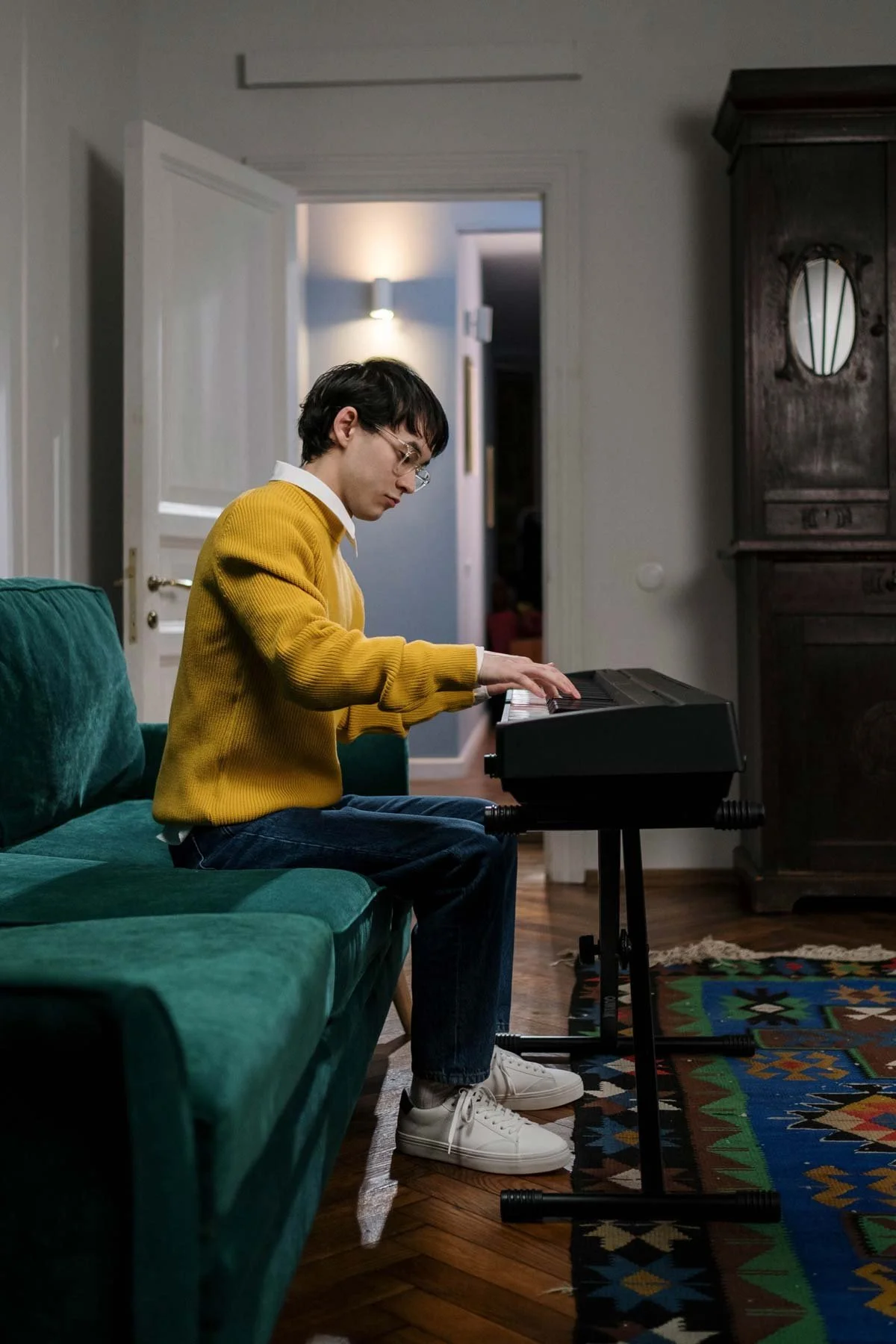 Side view of a person wearing a mustard-yellow sweater and glasses, sitting on a teal couch and playing an electronic keyboard