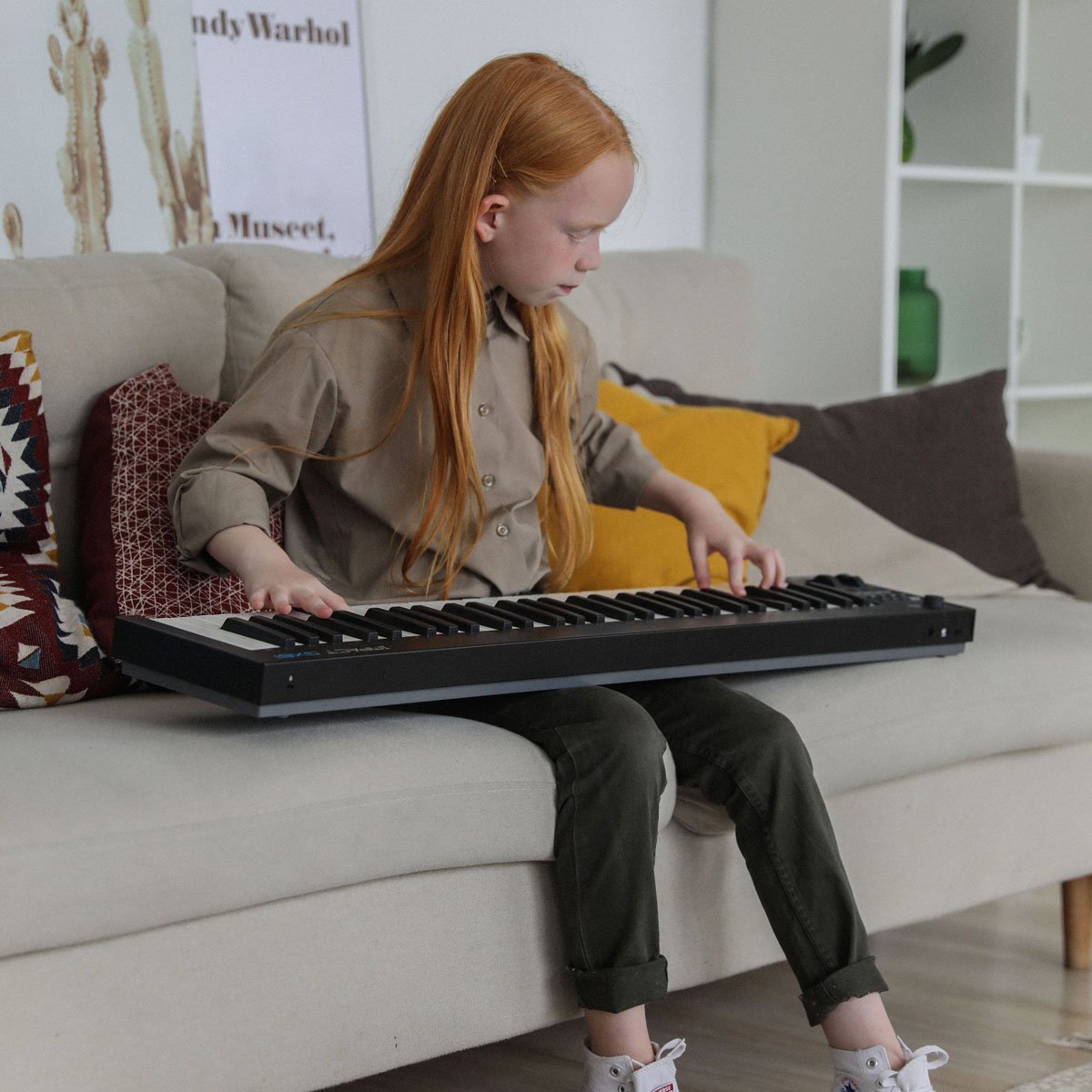 A pre-teen child with long red hair sits on a couch playing a keyboard on her lap.