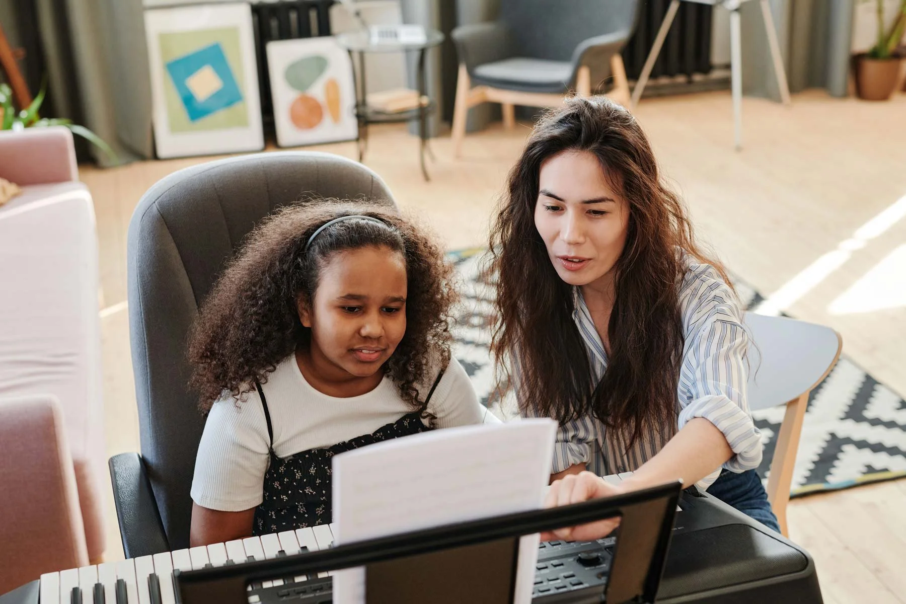 A young Black girl sitting at her keyboard reading sheet music with her teacher