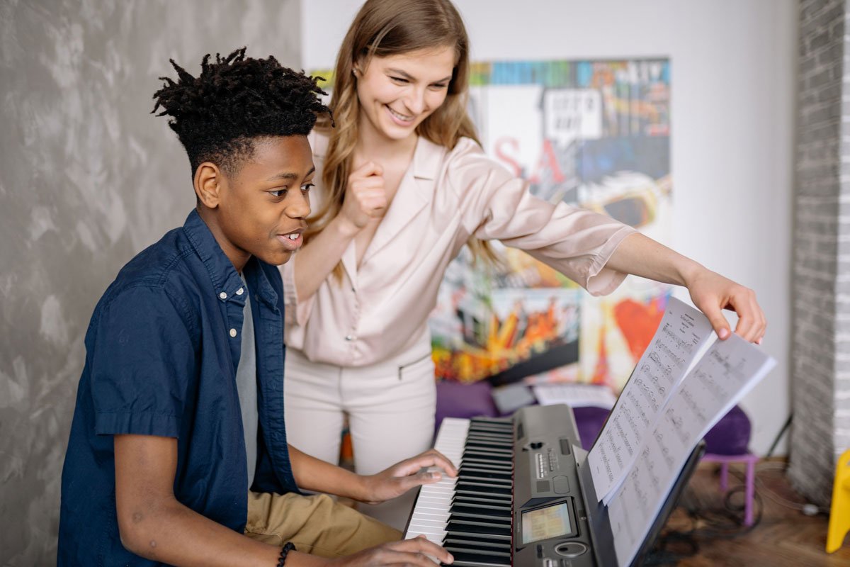 A music teacher smiles and points at sheet music while a Black teen boy plays a keyboard.