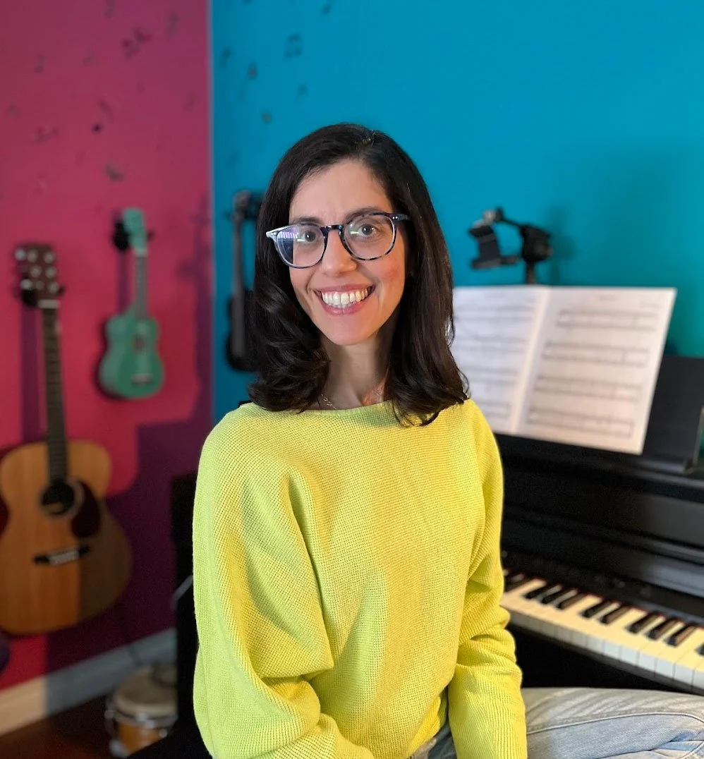 Nefeli sits in front of her piano and pink and teal walls with guitars and ukuleles hanging. She wears a sunny yellow top.