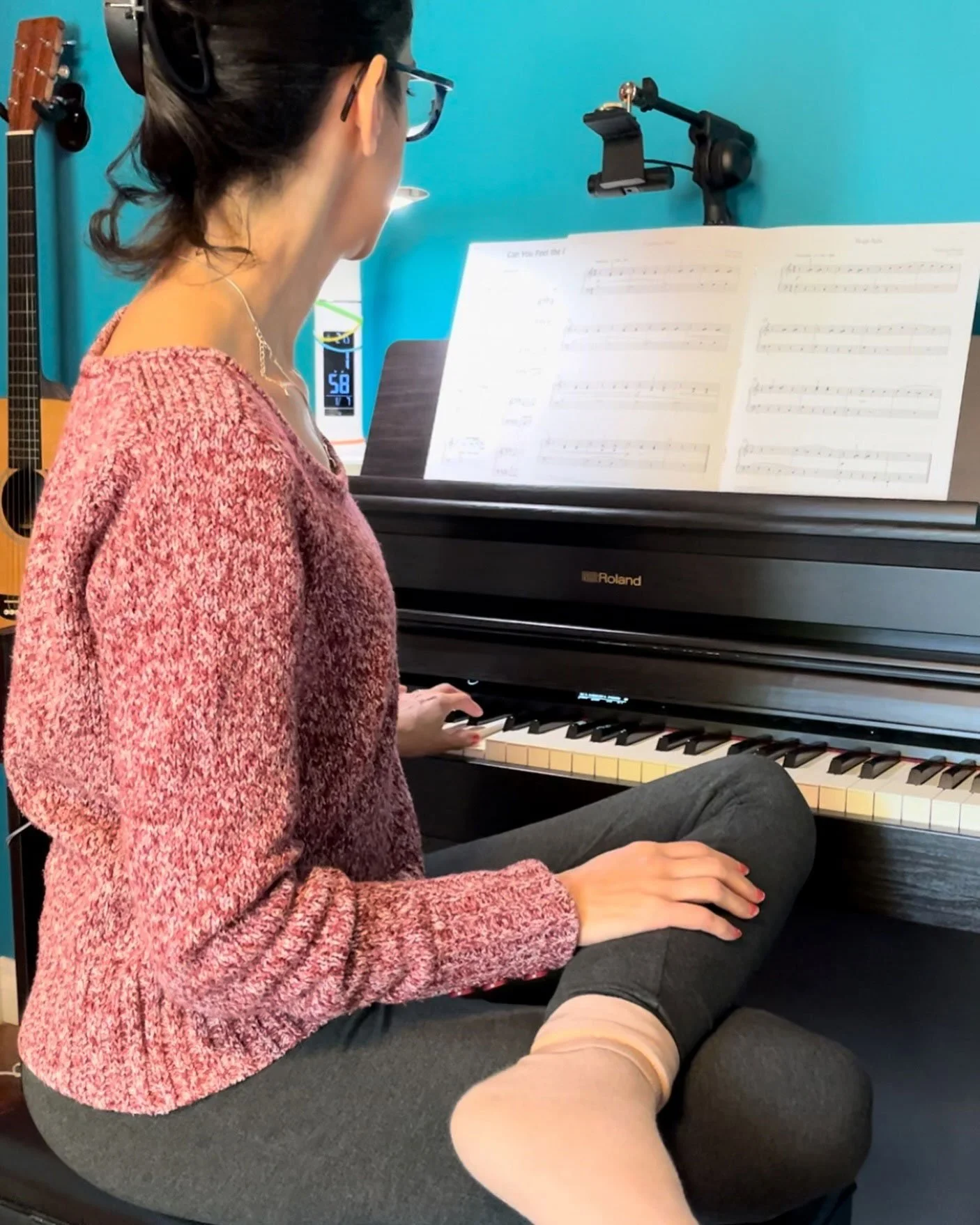 A side view of Nefeli sitting cross-legged on her piano stool, reading music with one hand on her piano and the other on her lap.