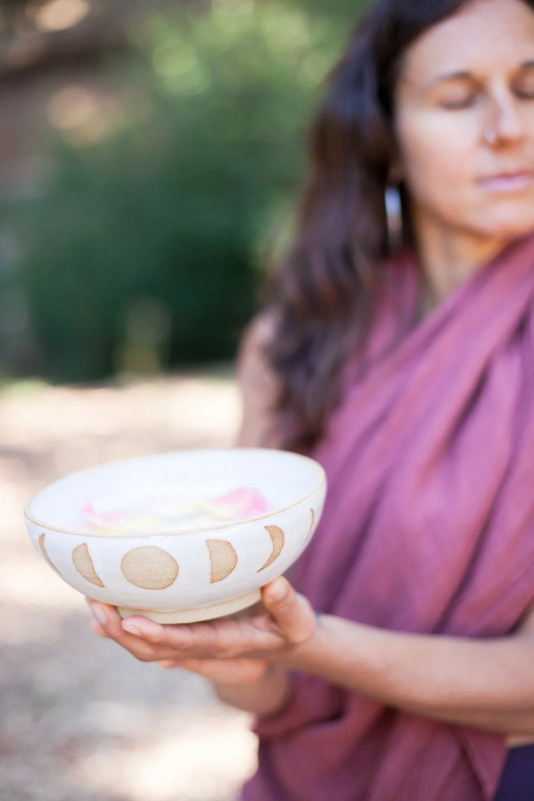A woman with long brown hair, wearing a purple shawl, holding a decorated ceramic bowl, outdoors with greenery in the background.