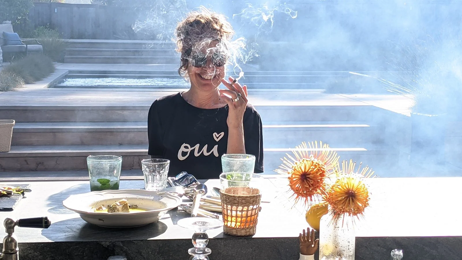 A woman with curly hair and glasses sitting at an outdoor table smoking a cigar. The table has plates, glasses, and a vase with orange flowers. There is a mist or smoke around her, and a pool with steps is visible in the background.