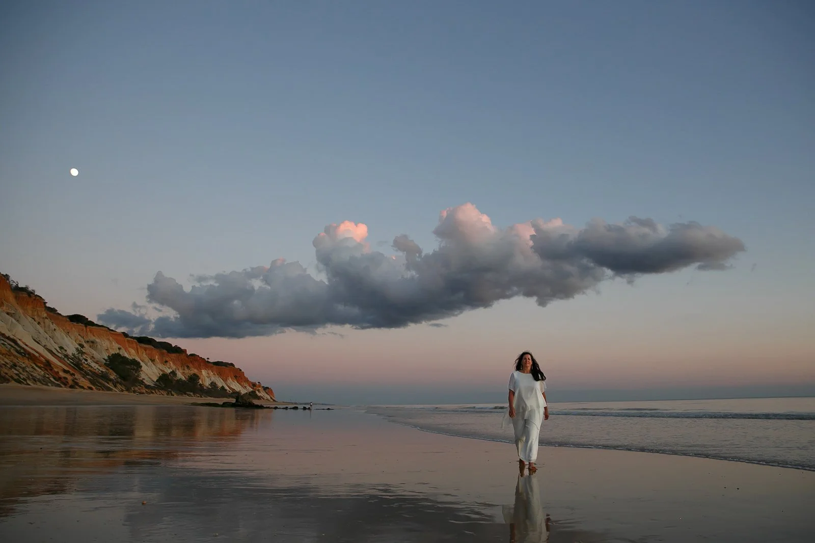 A woman in white walking along the shoreline of a beach at sunset, with a cliff on the left and clouds in the sky, and the moon visible in the sky.