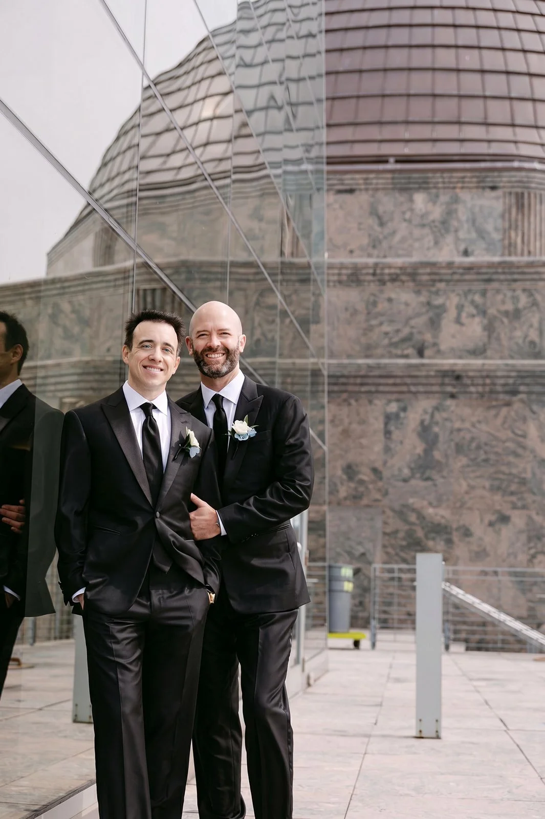 Two men in black tuxedos with white shirts and black ties, standing close together outside modern building with reflective glass and stone wall, smiling.