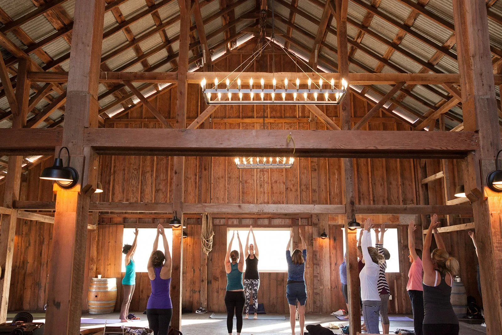 A group of people practicing yoga in a rustic wooden barn with large windows, wooden beams, and hanging chandeliers lighting the space.