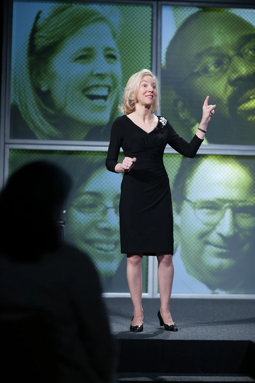 A woman with blonde hair, wearing a black dress and heels, giving a presentation on stage with three large screens behind her showing images of women with different facial features.