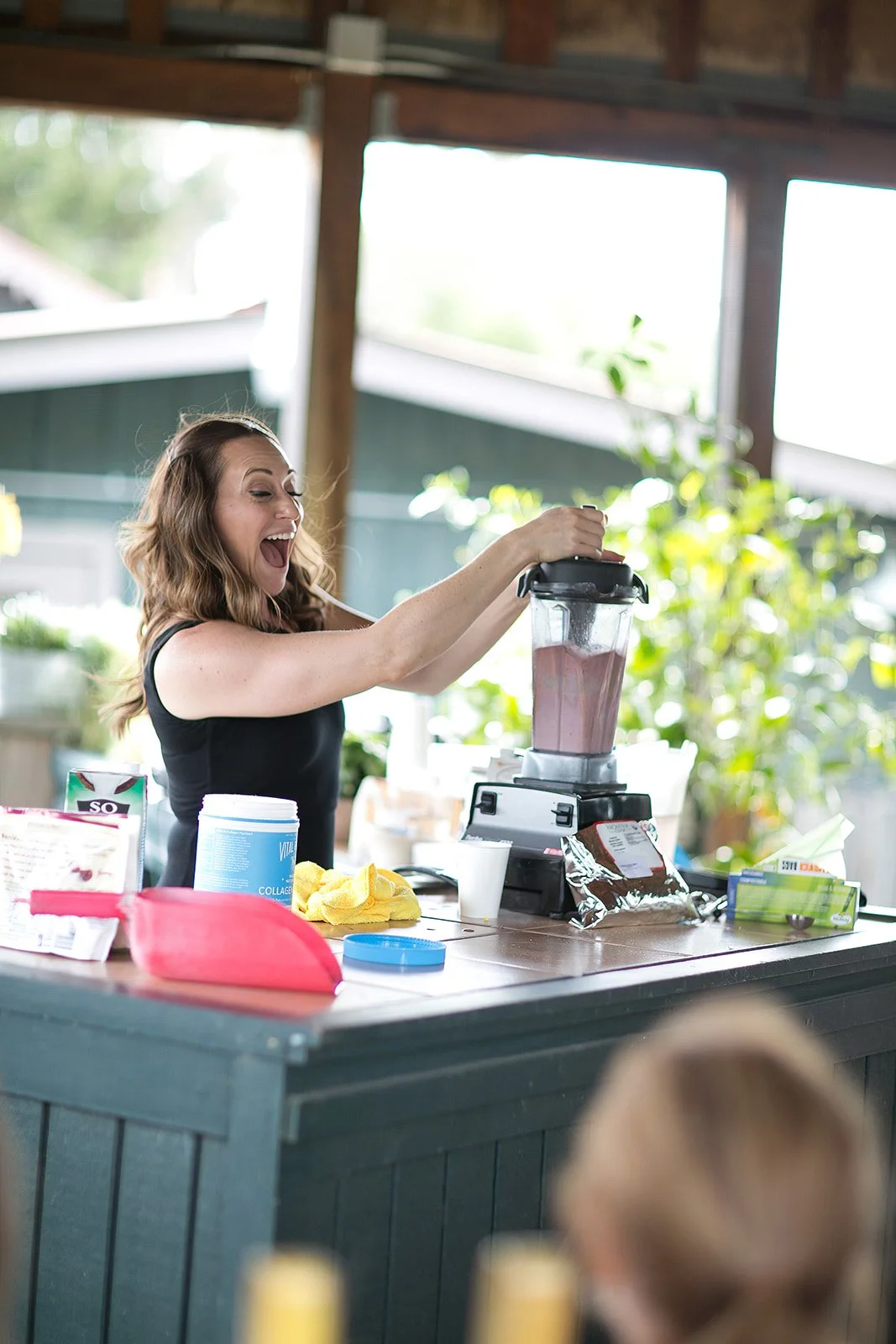 A woman making a pink smoothie in a blender at a kitchen counter, with various ingredients and utensils around.