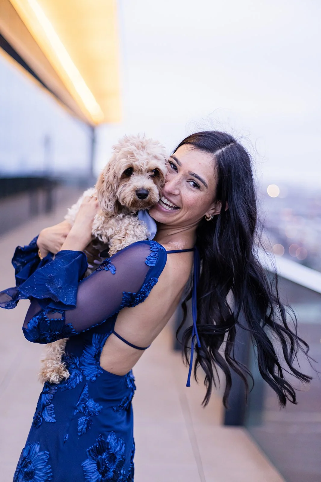 A woman with long dark hair wearing a blue dress holding a small curly-haired dog, smiling at the camera on a rooftop with city lights in the background.