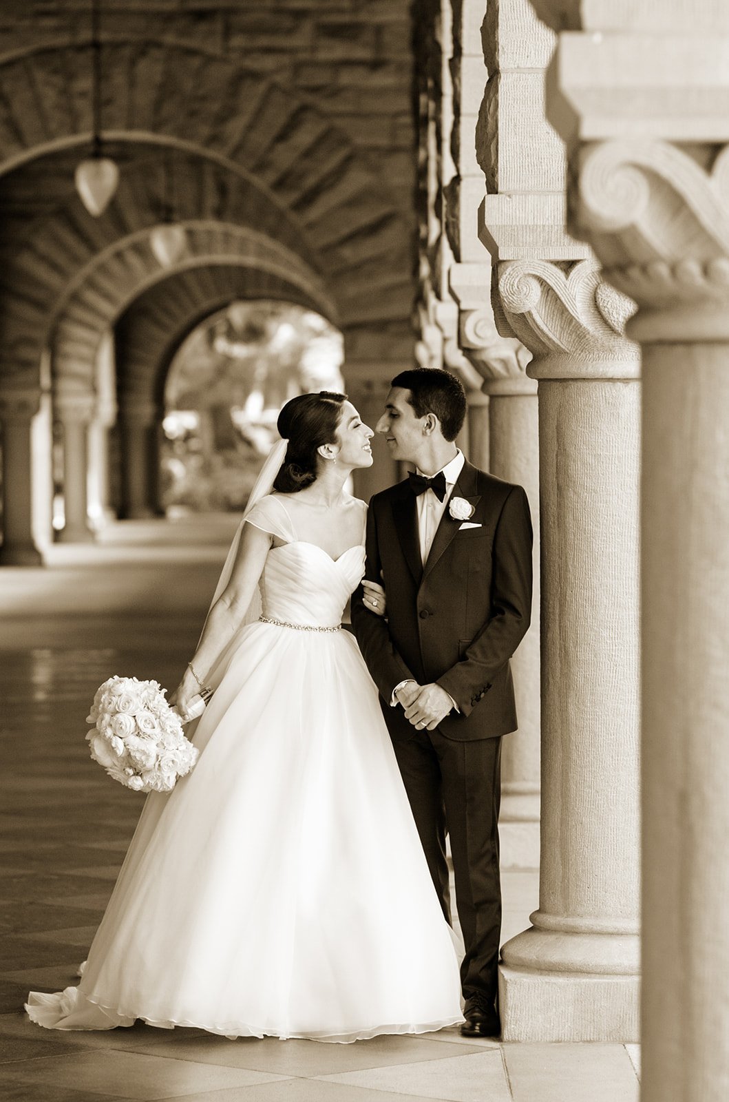 Black and white photograph of a bride and groom standing close together under an ornate stone archway, smiling and gazing into each other's eyes. The bride is holding a bouquet of flowers.