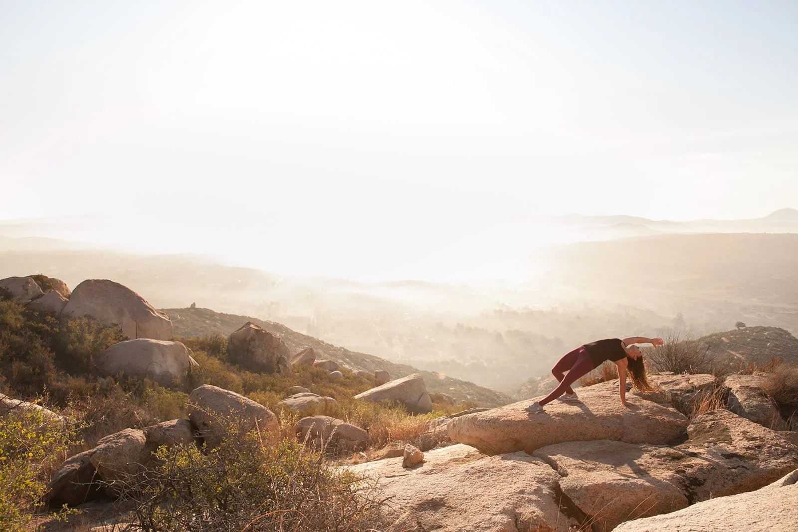 A person practicing yoga on a large rock on a mountain overlooking a valley at sunrise.