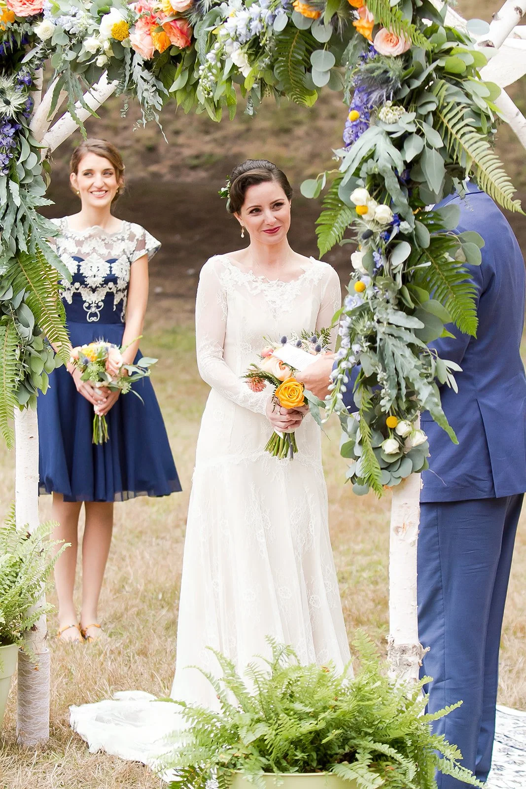 A wedding ceremony outdoors with a bride and groom, two women in dresses, and a floral arch with green leaves and colorful flowers.