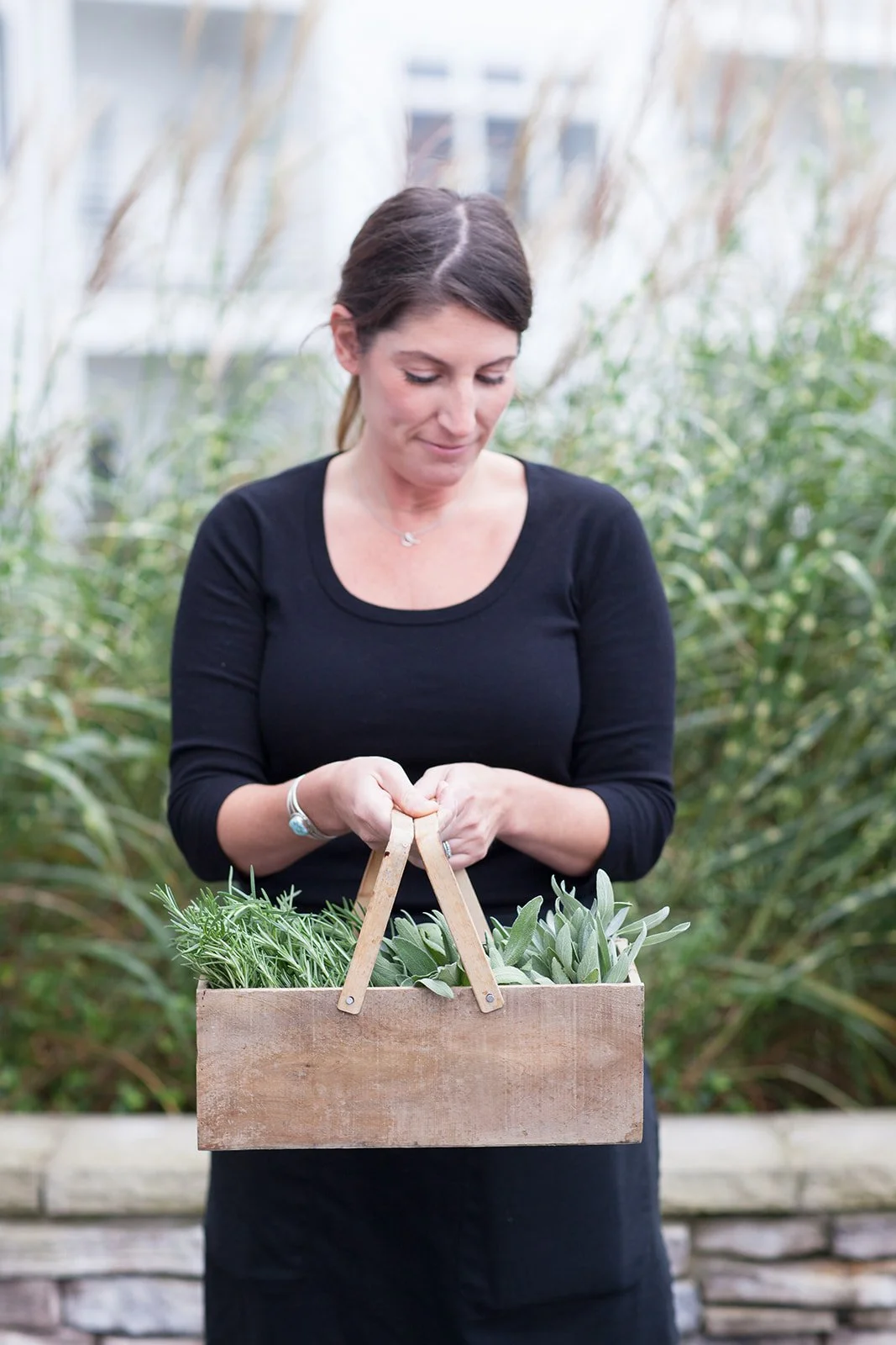 A woman in a black top holding a wooden basket filled with herbs in a garden