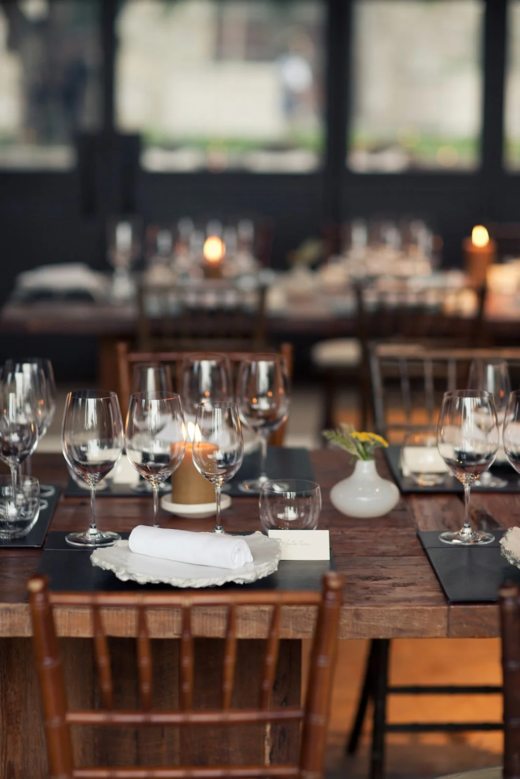 Table setting for a fine dining restaurant, with wine glasses, white napkin, small vase with yellow flowers, candle, and place card on a wooden table.