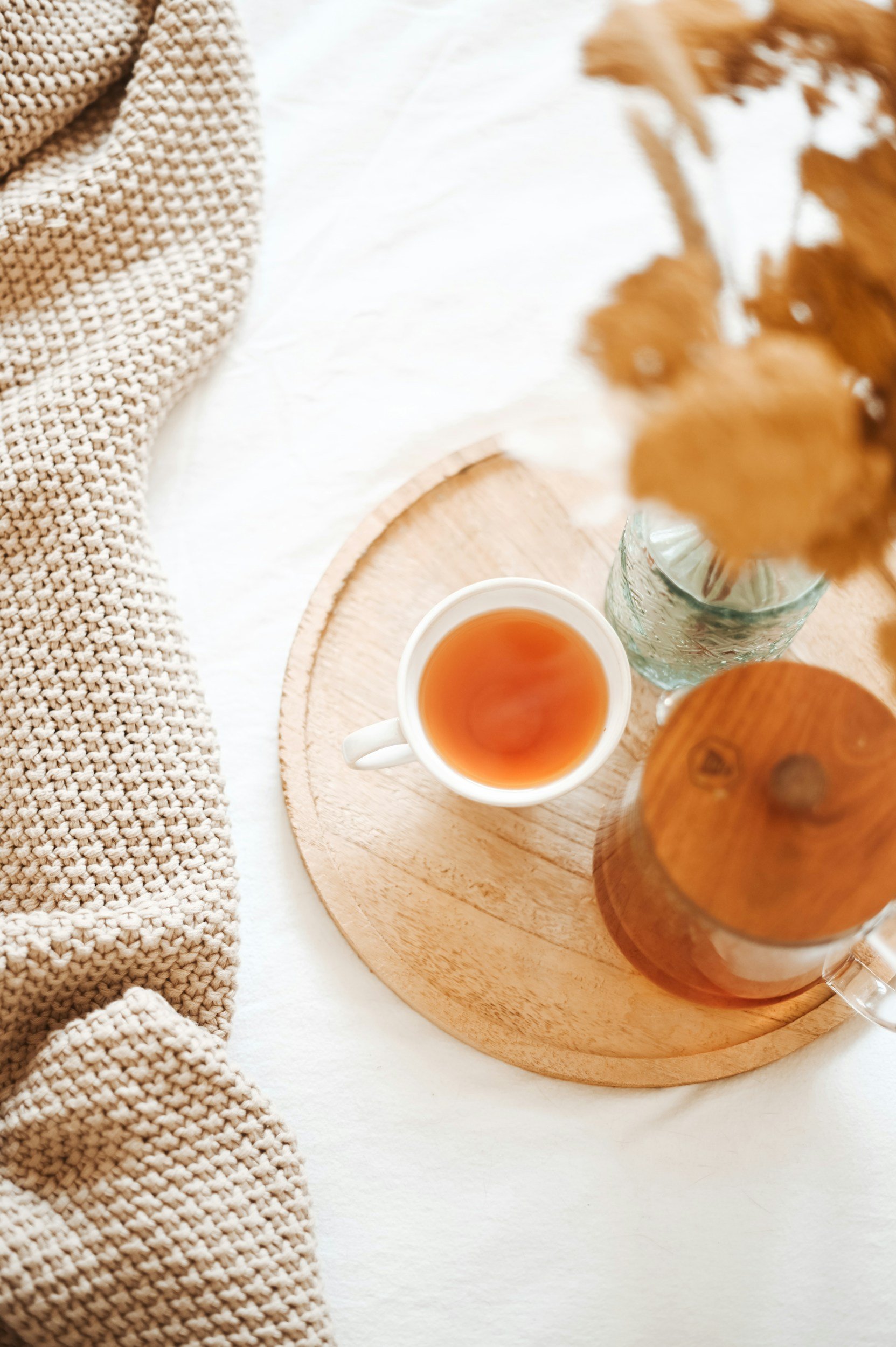 A white cup of tea on a wooden round tray next to a glass of water, with a beige knitted blanket on the left and a bouquet of dried brown leaves on the right.