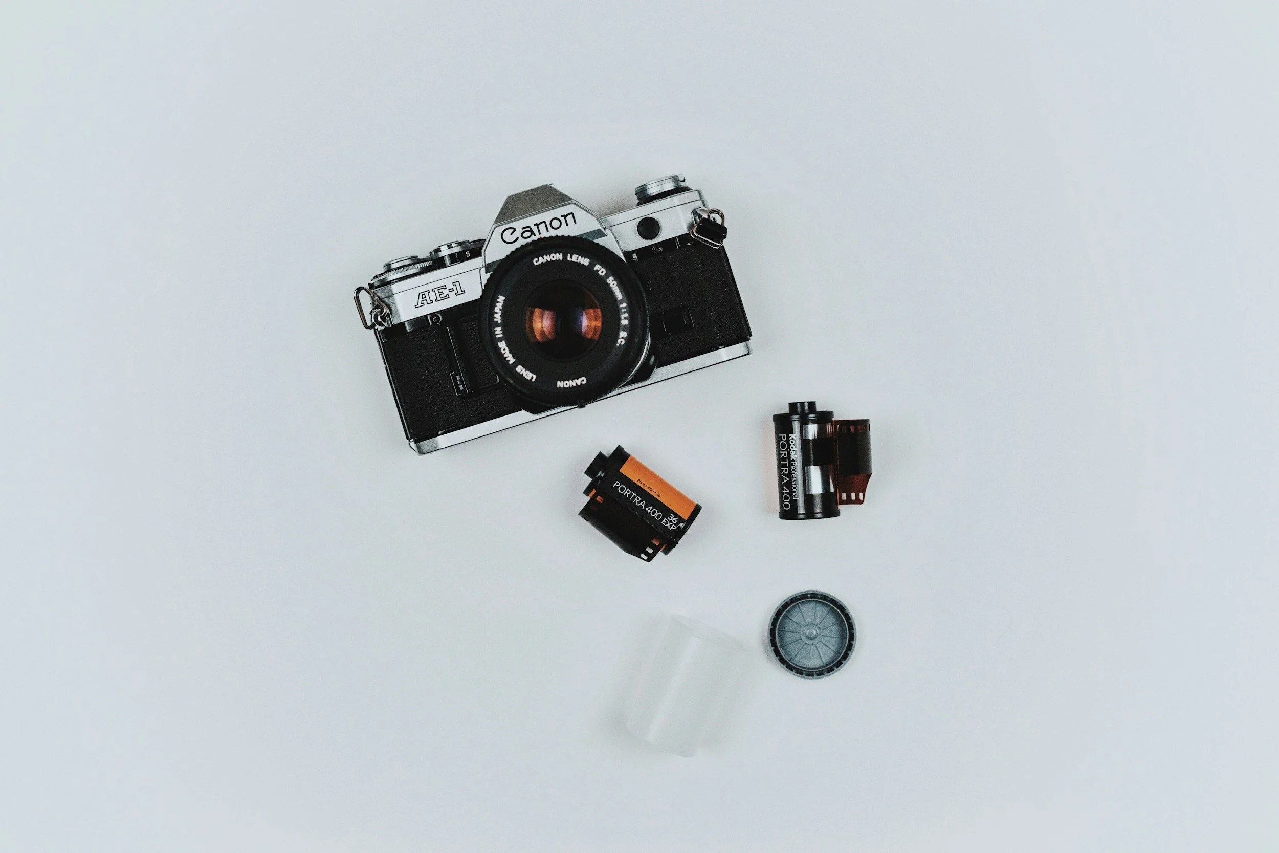 A vintage black and silver Canon AE-1 film camera with an attached lens is on a white surface, surrounded by film rolls, a lens cap, and film canisters.