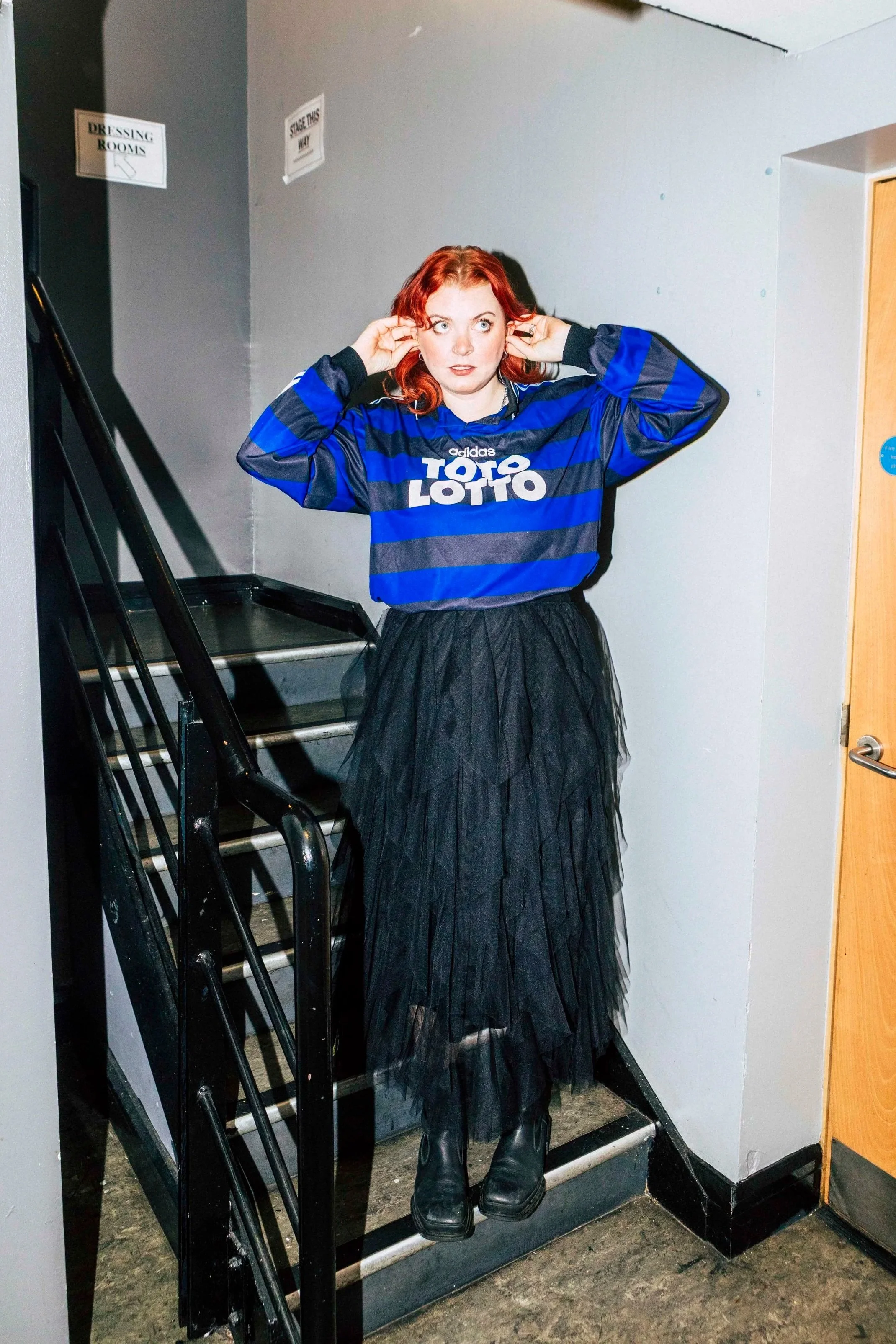 A woman with red hair wearing a blue and black sports jersey and a black tulle skirt standing on a staircase in an indoor setting.