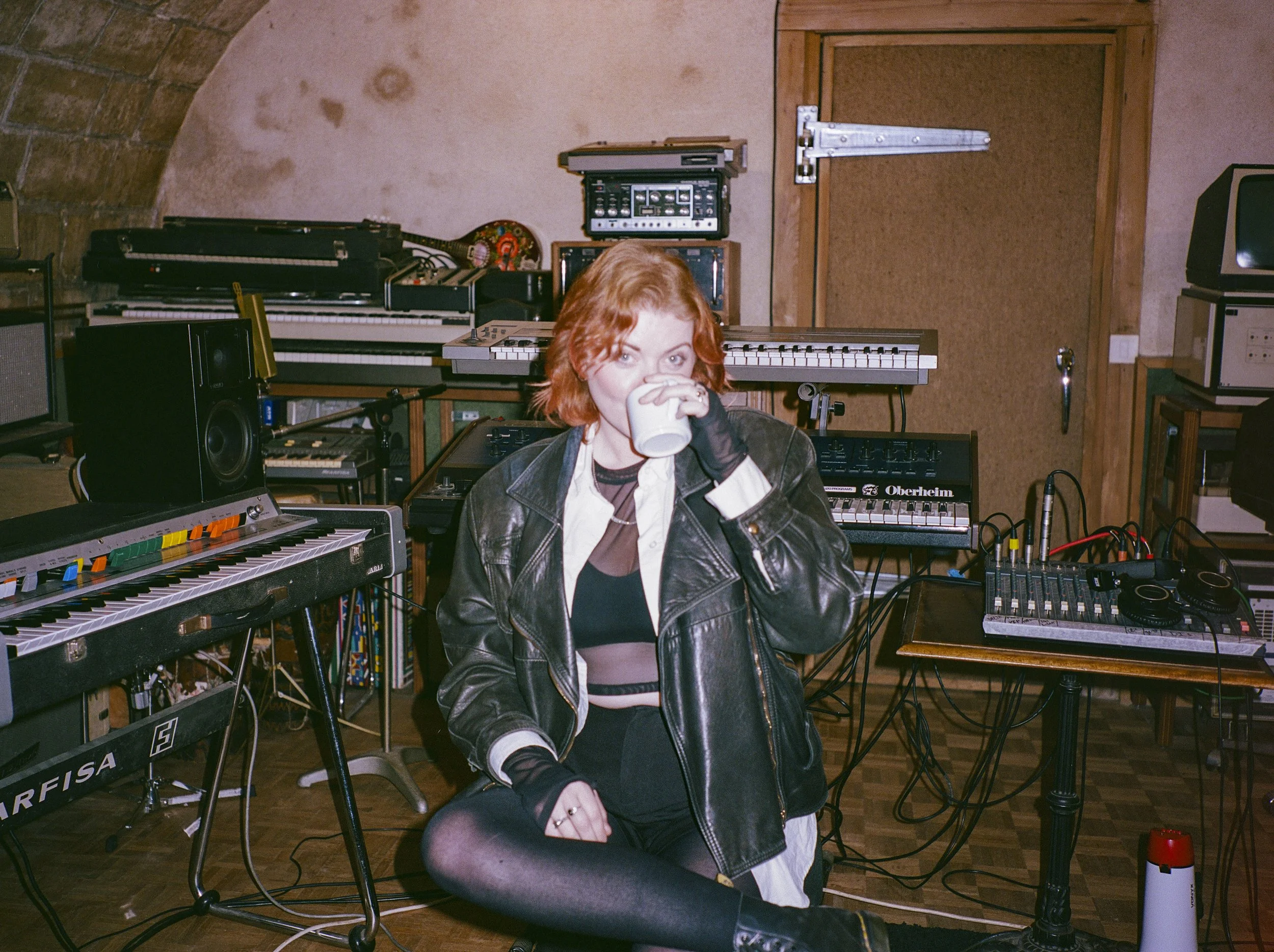 A young woman with red hair drinking from a white mug in a music studio filled with keyboards, synthesizers, speakers, and electronic equipment, with a woman wearing a black leather jacket and striped shirt sitting nearby.