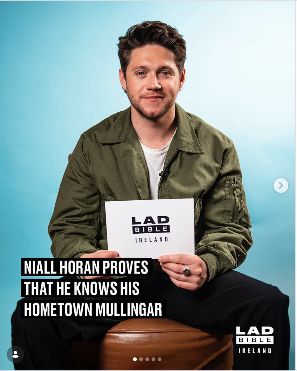 A young man with brown hair and a beard, wearing a green jacket and white t-shirt, sitting on a brown stool against a blue background, holding a card that says "LAD Bible Ireland."