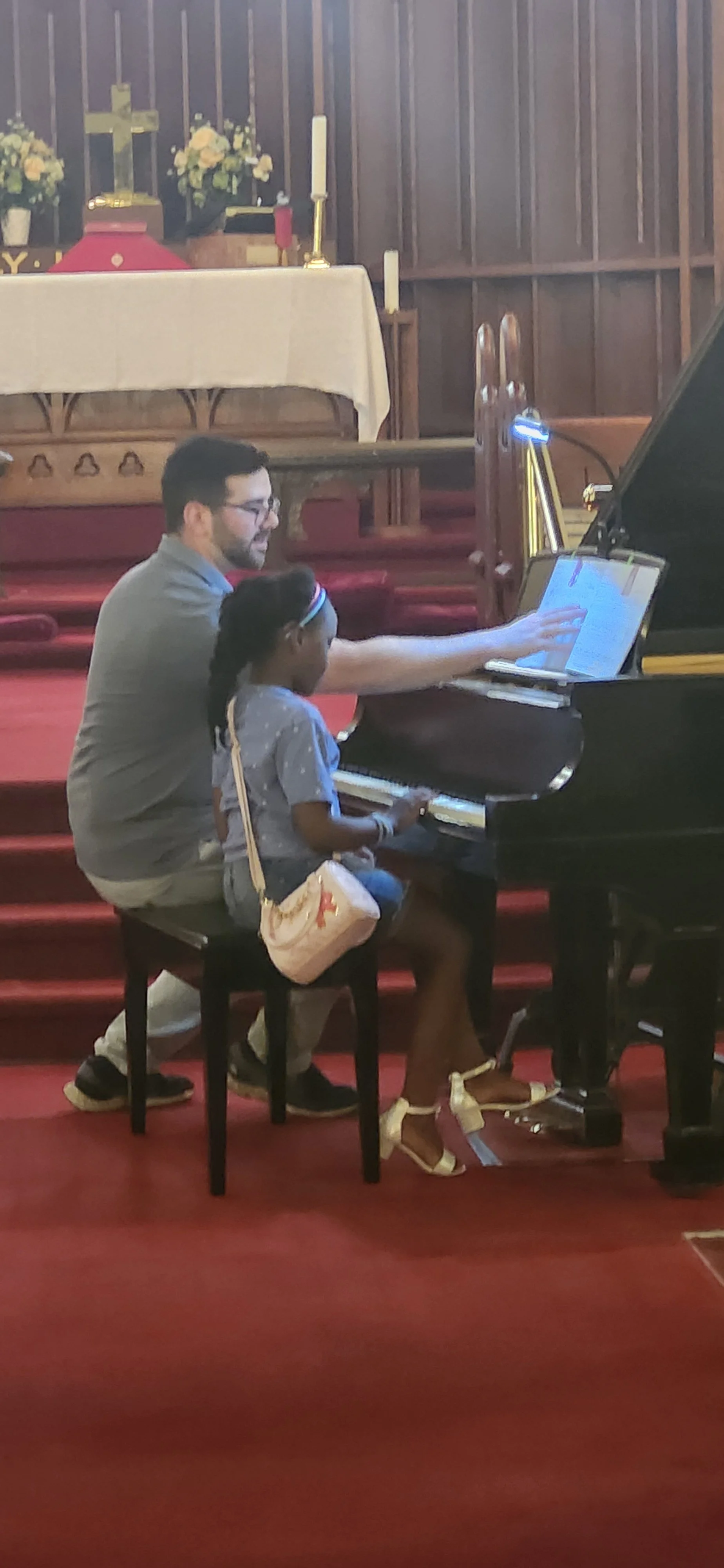 A man and a young girl playing piano together inside a church with a religious altar in the background.