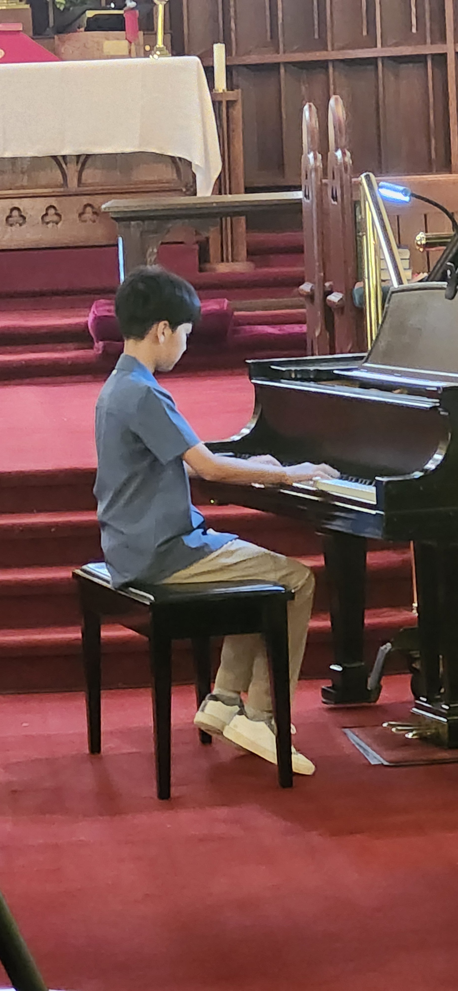 A young boy sitting on a black piano bench playing a grand piano inside a church with red carpeted stairs and an altar in the background.