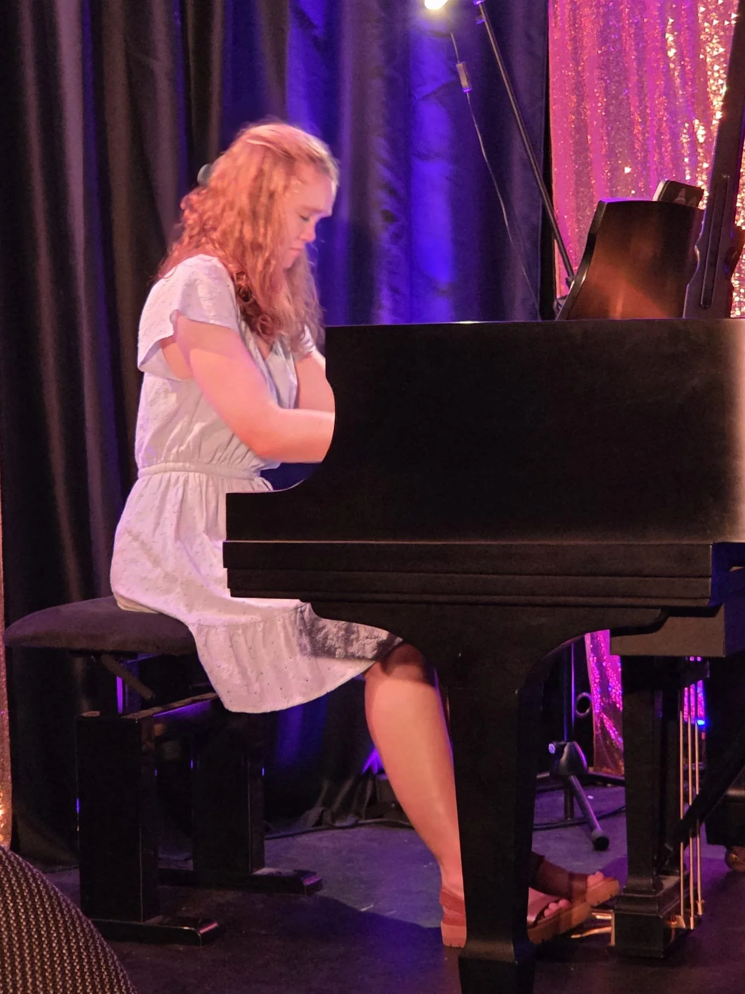 A young girl with curly red hair playing a black grand piano on a stage, with dark curtains and purple lighting in the background.