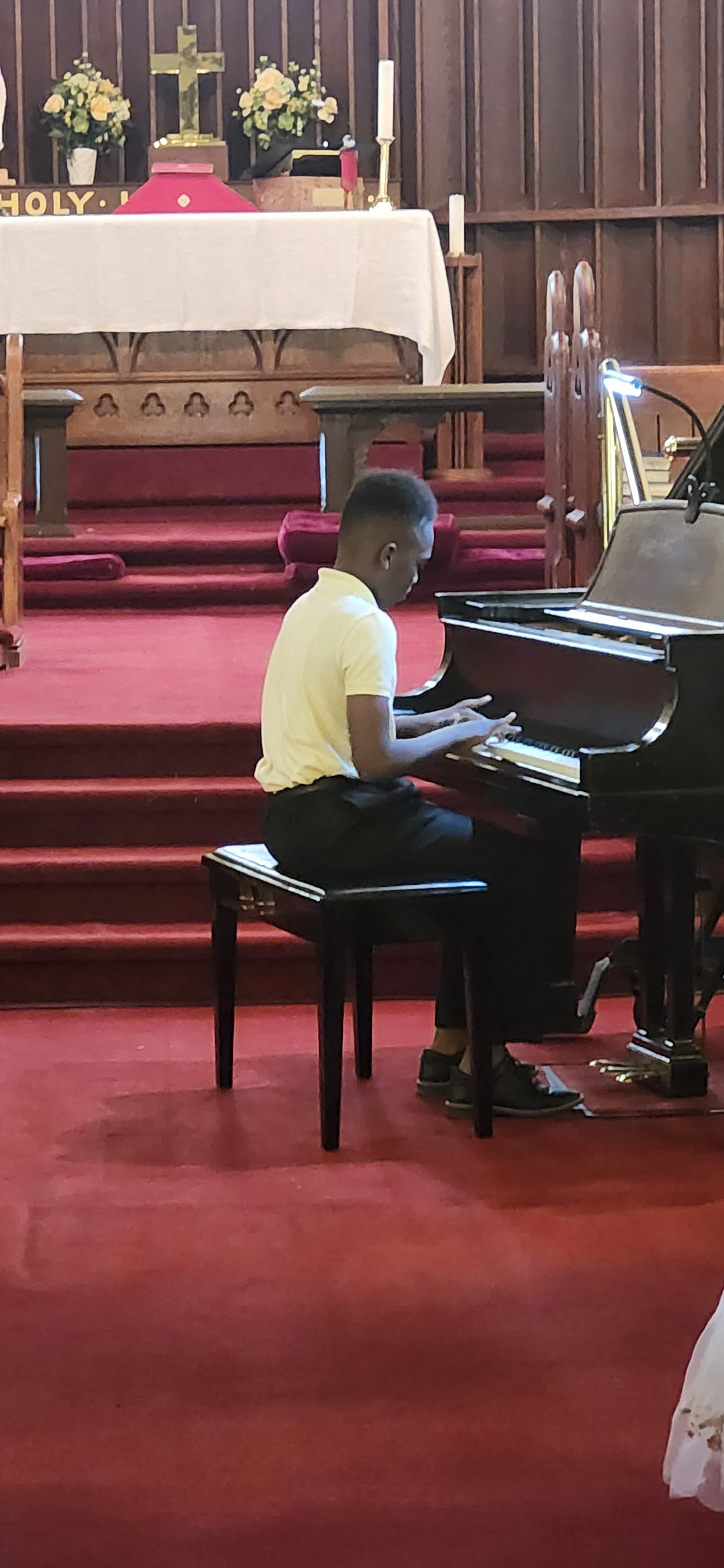 A young boy playing a grand piano in a church or chapel with red carpeted steps and religious decorations on the altar, including flowers, a crucifix, and candles.