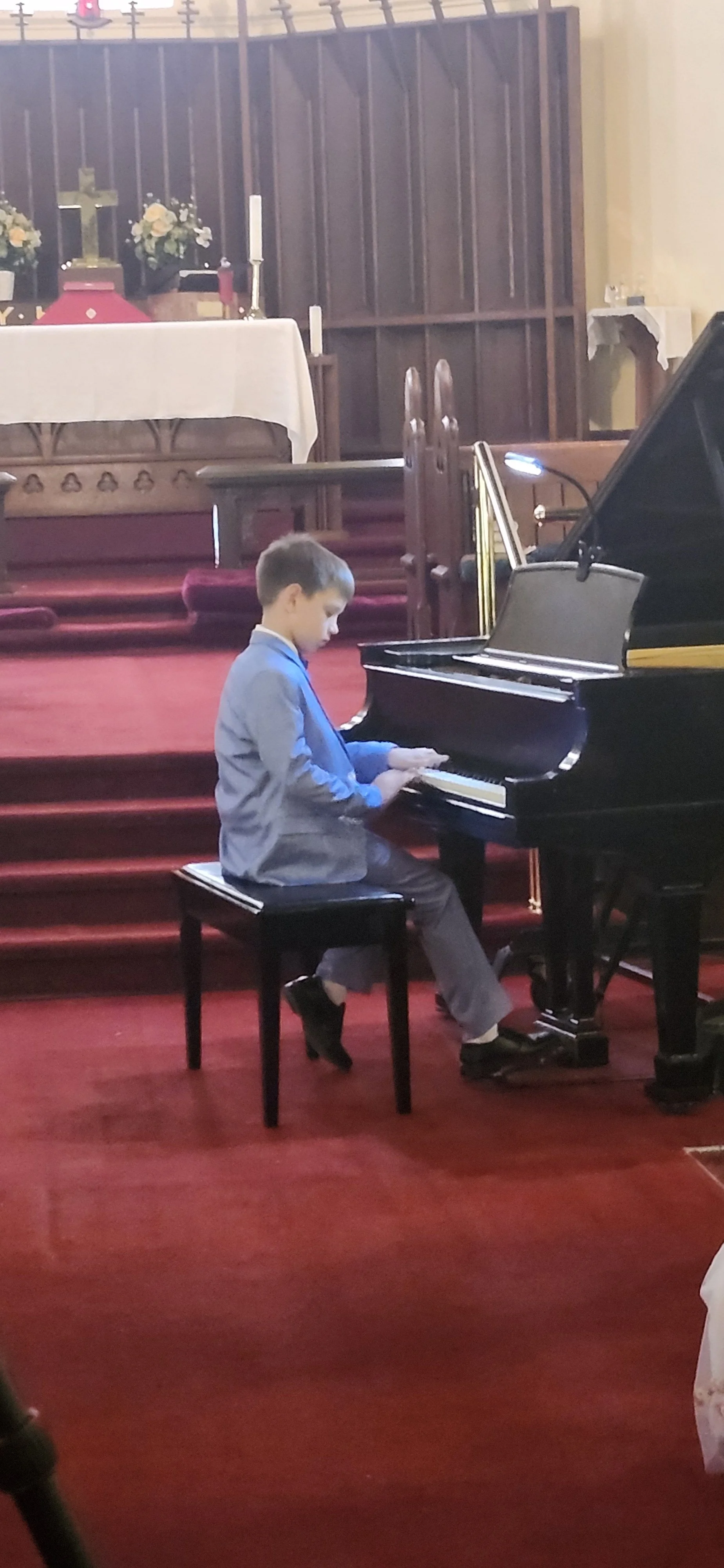 A young boy in a gray suit is sitting on a black bench, playing a grand piano in a church setting with an altar and religious decorations in the background.