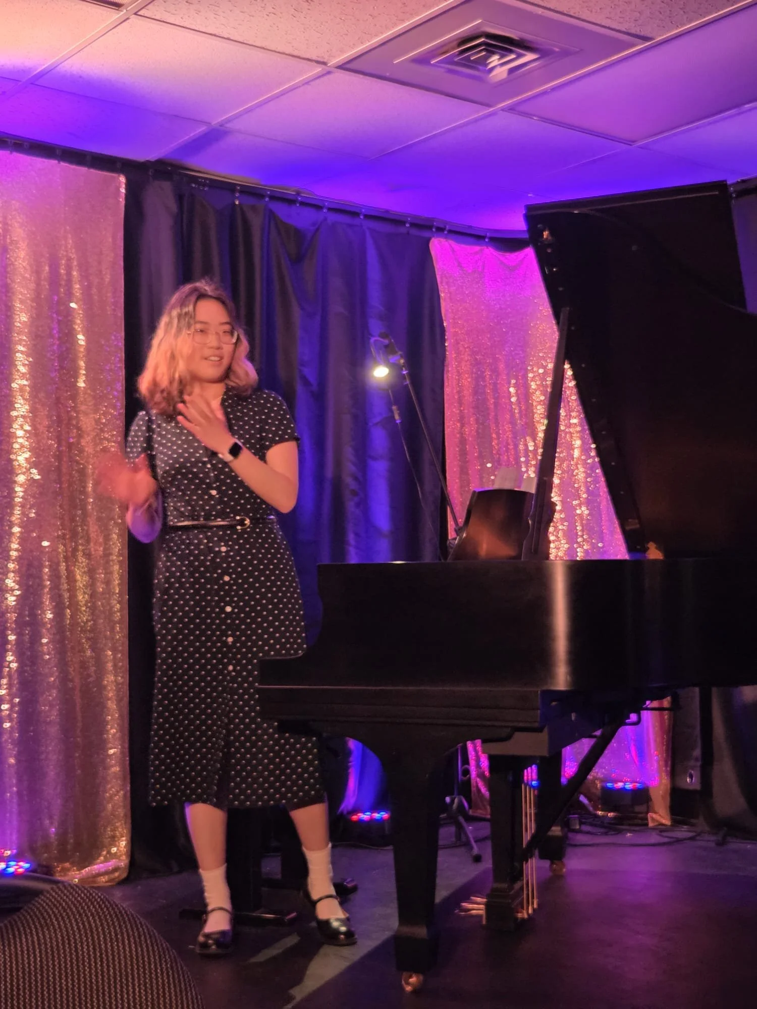 A woman with wavy hair and glasses in a polka dot dress standing on a stage with a black grand piano, black curtain, and purple stage lighting.