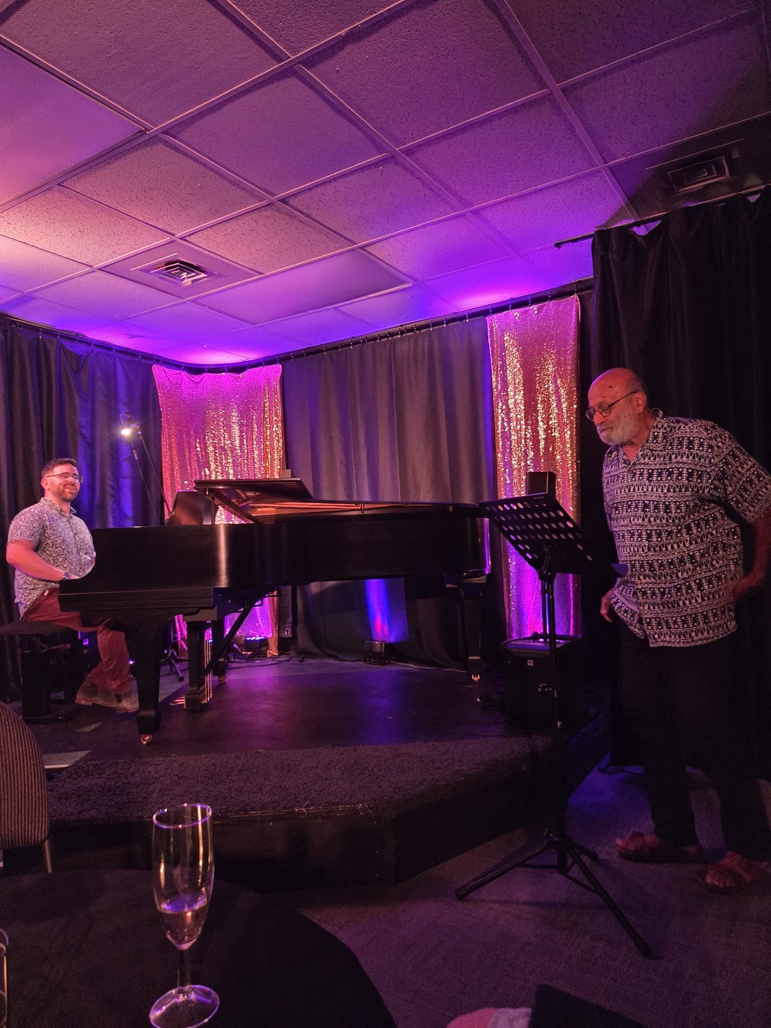 Two Elli Kozak music school students in a dimly lit room with purple lighting, a grand piano, dark curtains, and sparkling pink decorations in the background.