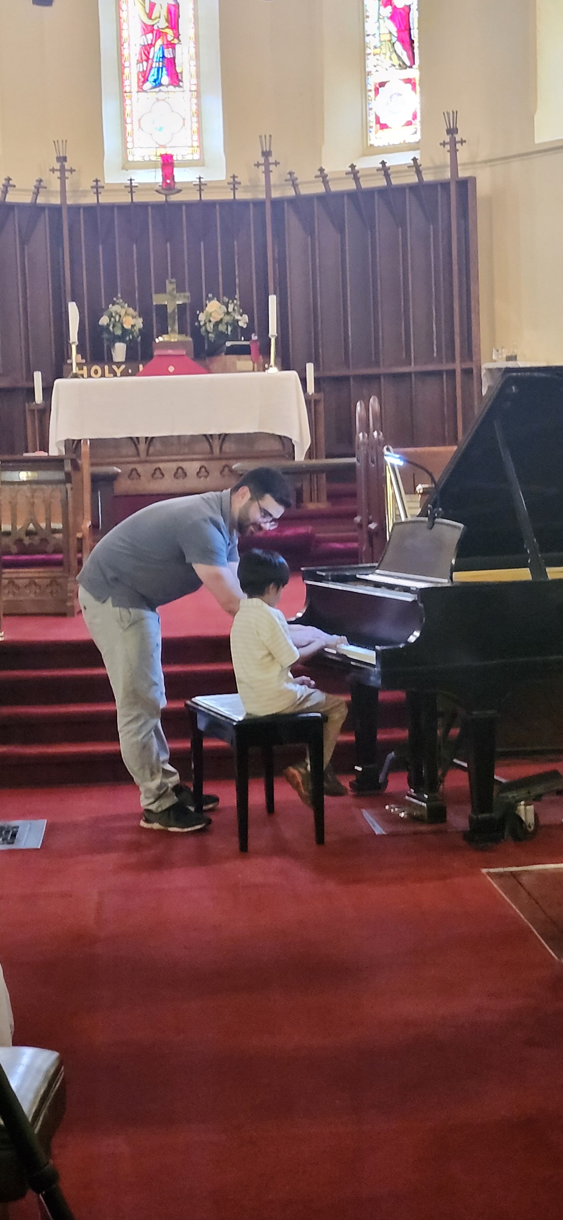 A man helps a young boy play the piano inside a church with stained glass windows and religious decor.