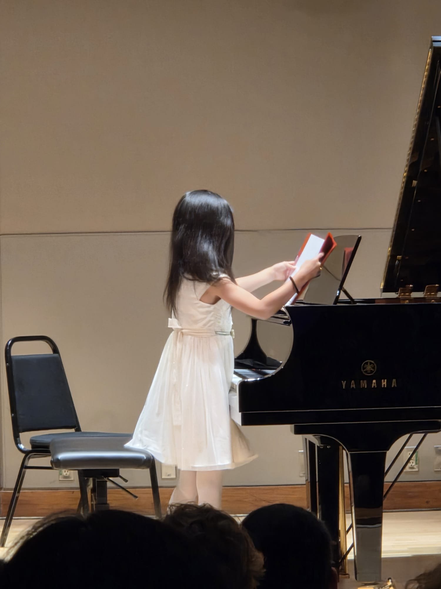 A girl with long dark hair wearing a white dress is playing a Yamaha grand piano on a stage, with chairs in the background and audience members visible at the bottom of the image.