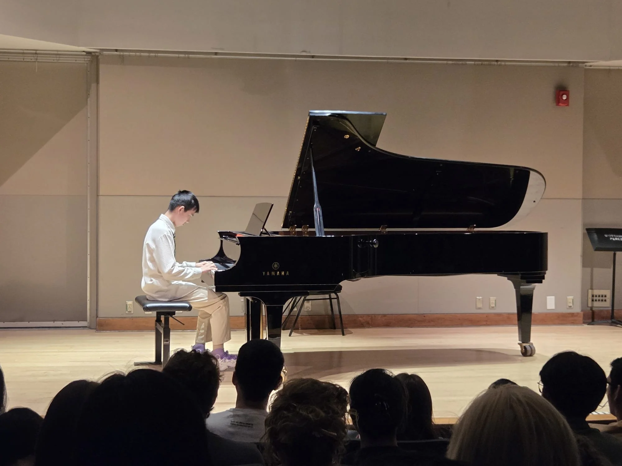 A young person wearing a white shirt and beige pants playing a black Yamaha grand piano on a stage, with an audience watching.