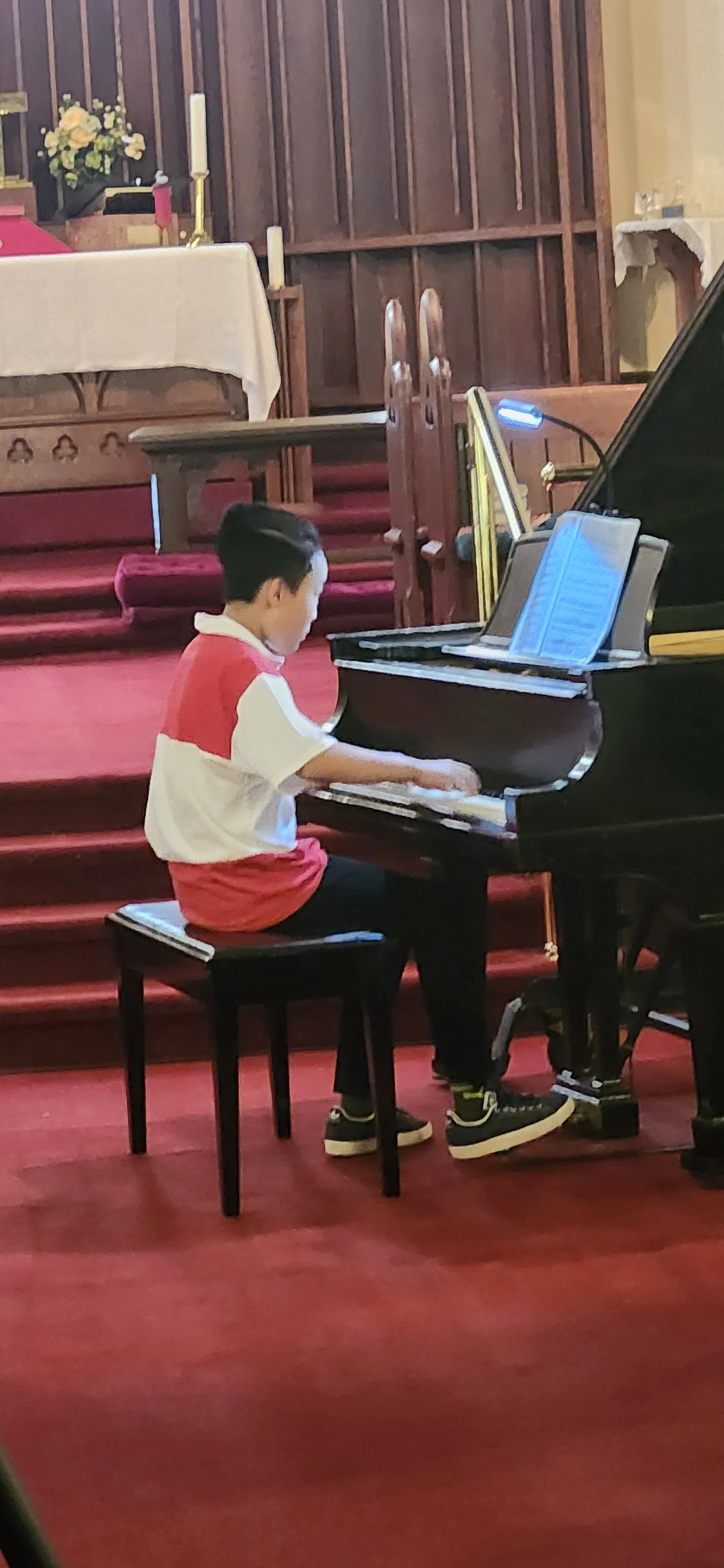 A boy in a red and white shirt playing a grand piano in a church or chapel, sitting on a stool, with a lit altar and wooden pews in the background.