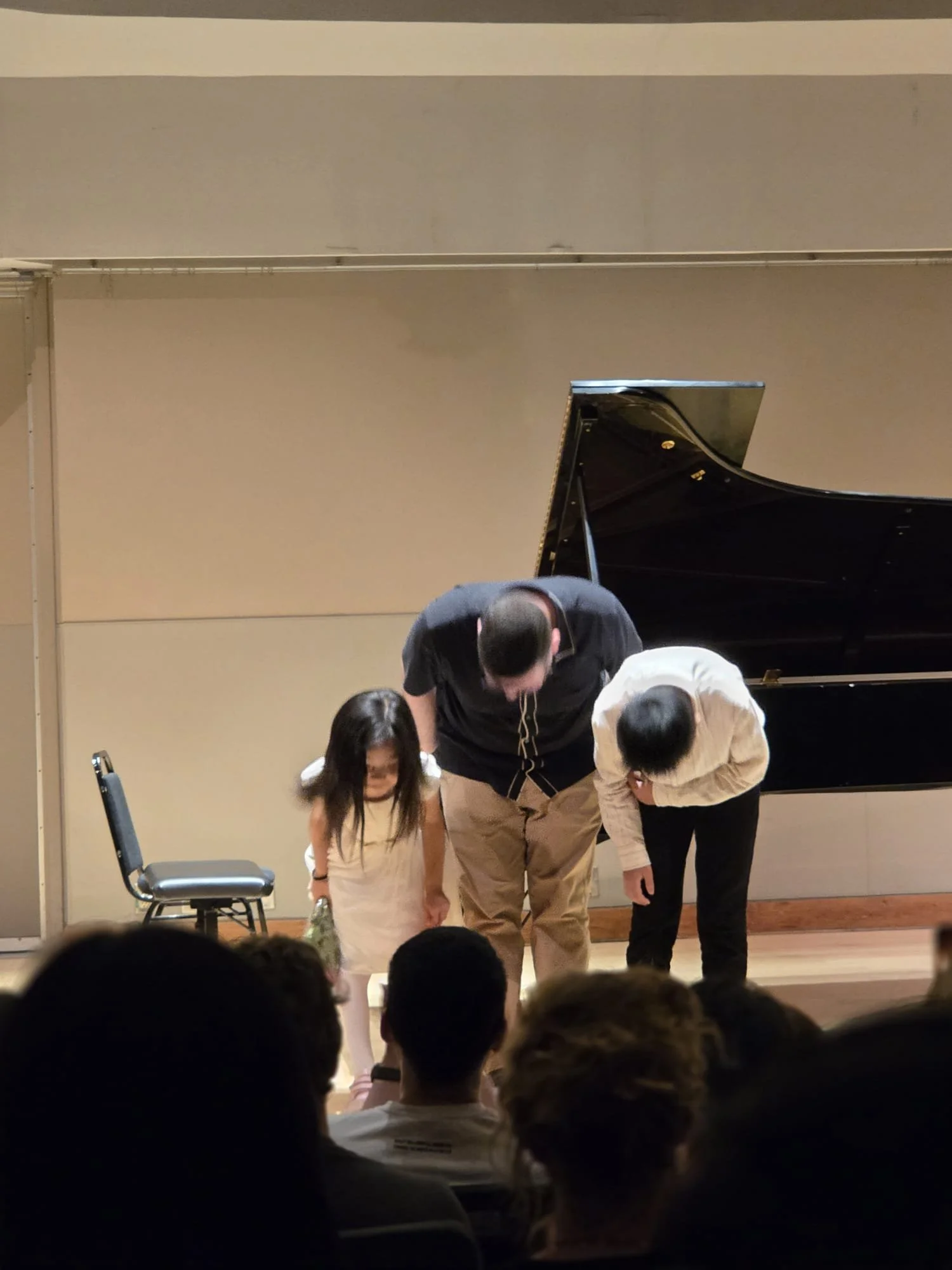 A pianist bows with three children on stage after a performance, with an audience in front.