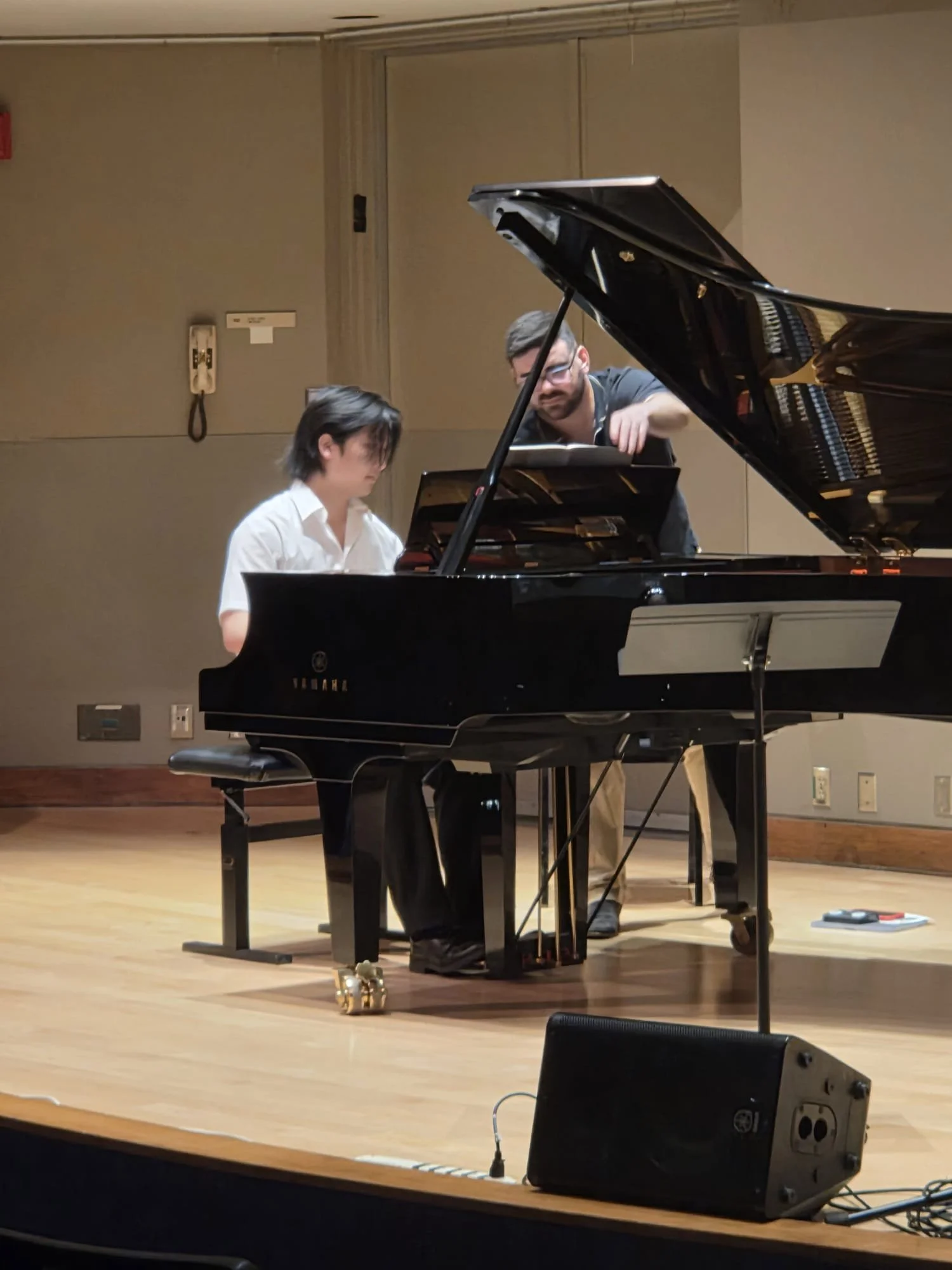A person with dark hair and glasses sits at a grand piano, playing or practicing, while another person with dark hair, glasses, and a beard looks on, possibly instructing or assisting, in a music rehearsal space with wooden floors and beige walls.