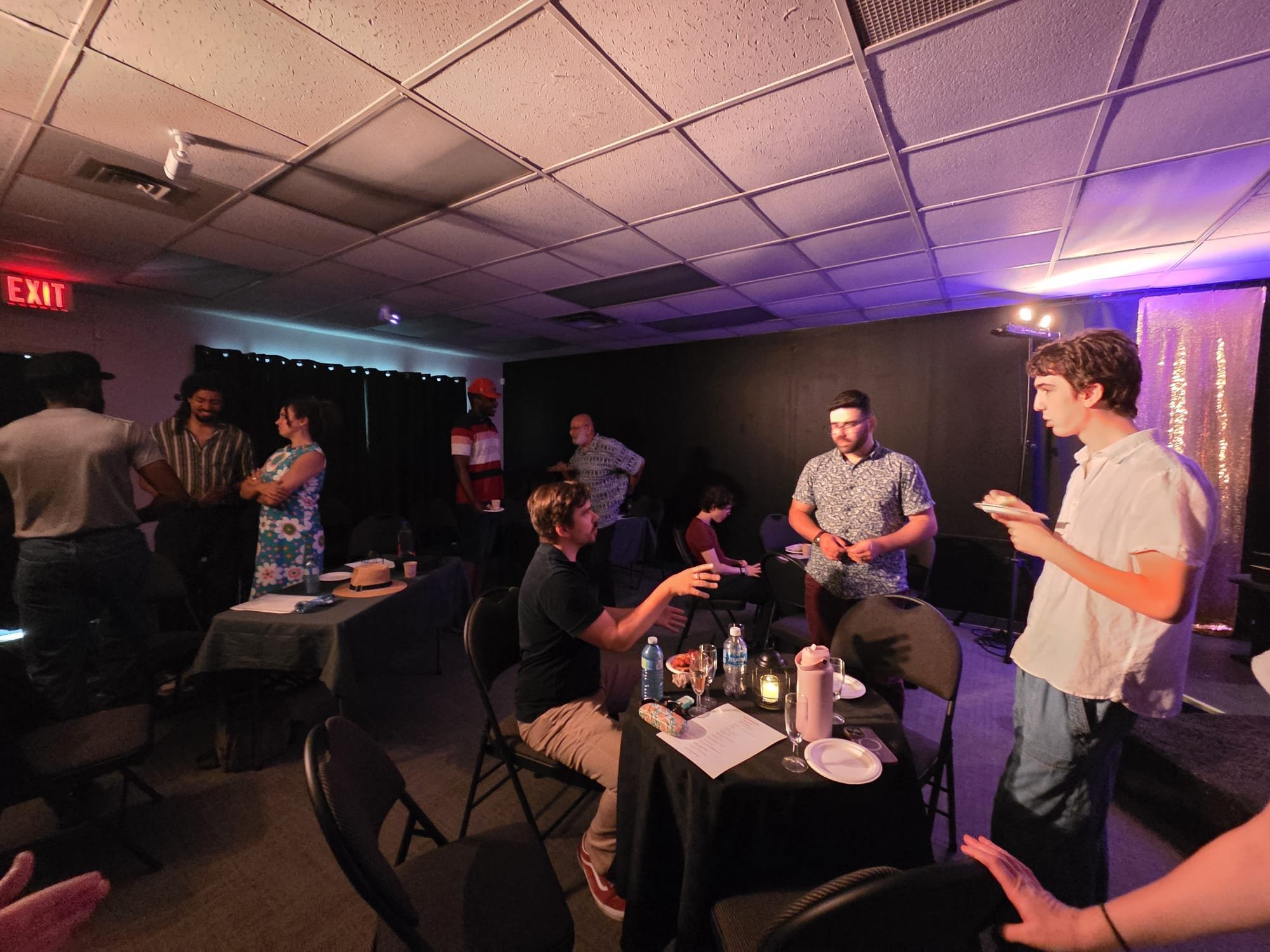 People gather in a dimly lit room with tables, chairs, and drinks, engaged in conversations; a young man in a white shirt stands reading from a paper, others stand and sit around, with some watching him. The ceiling has purple and blue lighting, and 