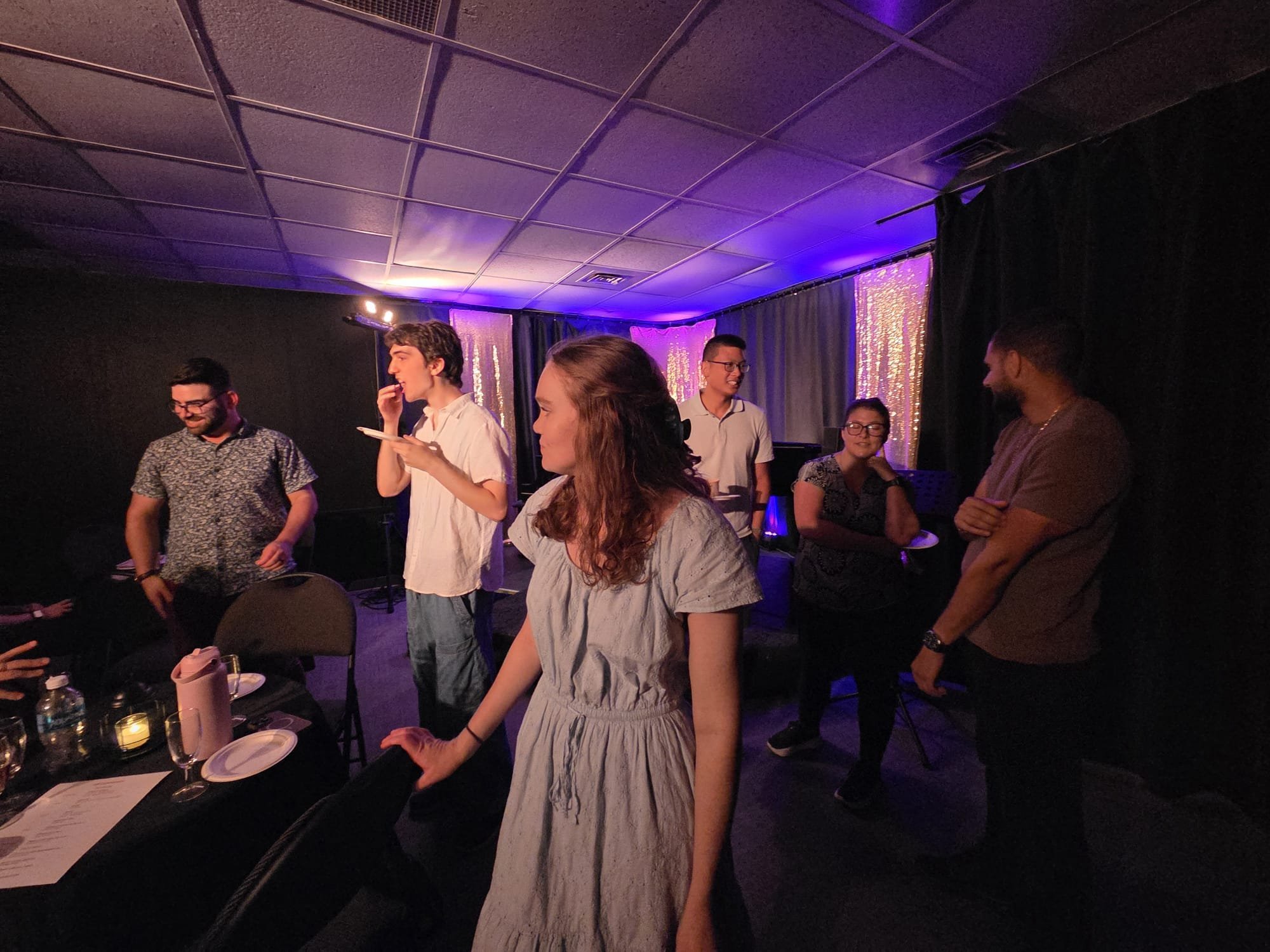 A group of people gathered in a dimly lit room with purple lighting, some standing and some sitting at tables with drinks and plates, possibly at a social event or party.