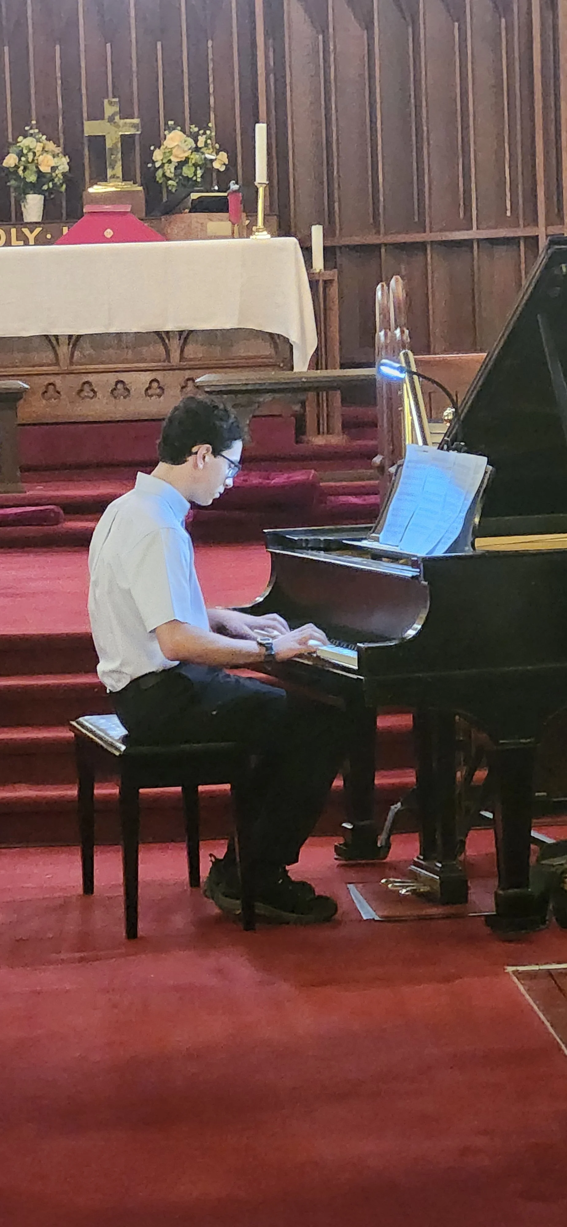 A young man in a white shirt playing a black grand piano in a church with a red carpet, floral arrangements, a cross, and candles in the background.