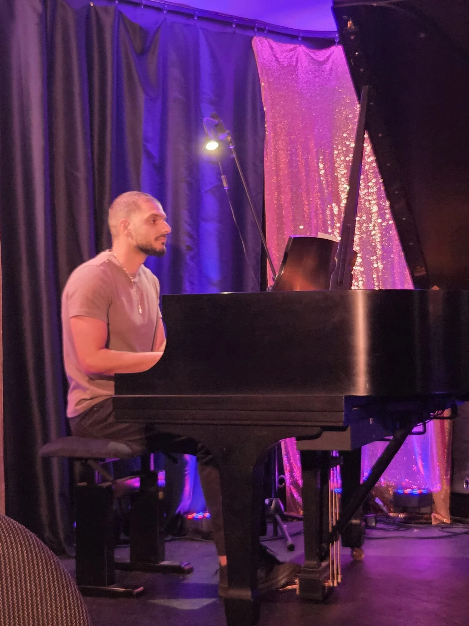 A man with short blond hair and a beard sitting at a black grand piano on a stage with purple and pink lights, black curtains, and a sparkling pink backdrop.