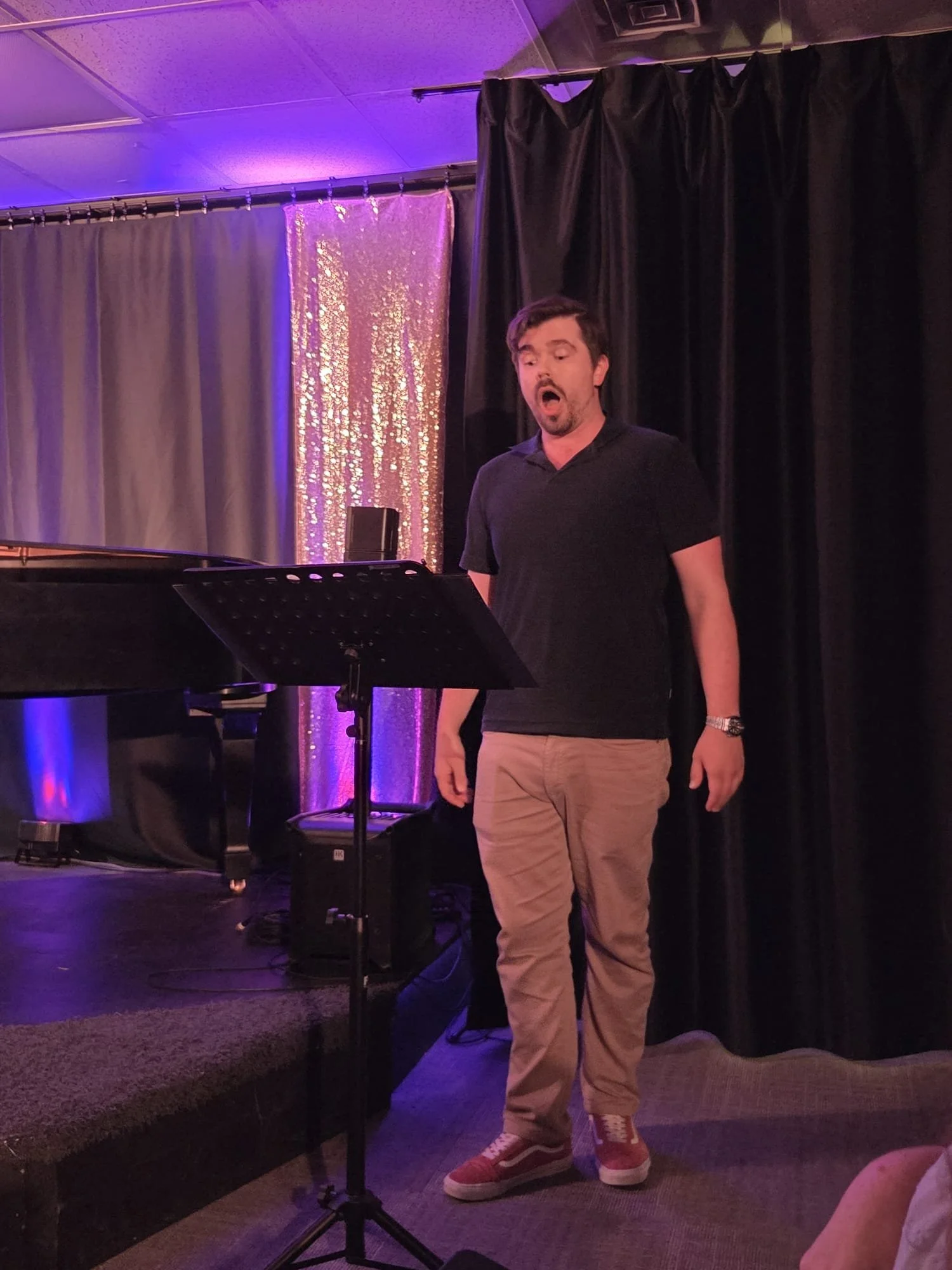 A man singing or performing on stage with a microphone stand, black curtains behind him, purple and black lighting, and a piano to his left.