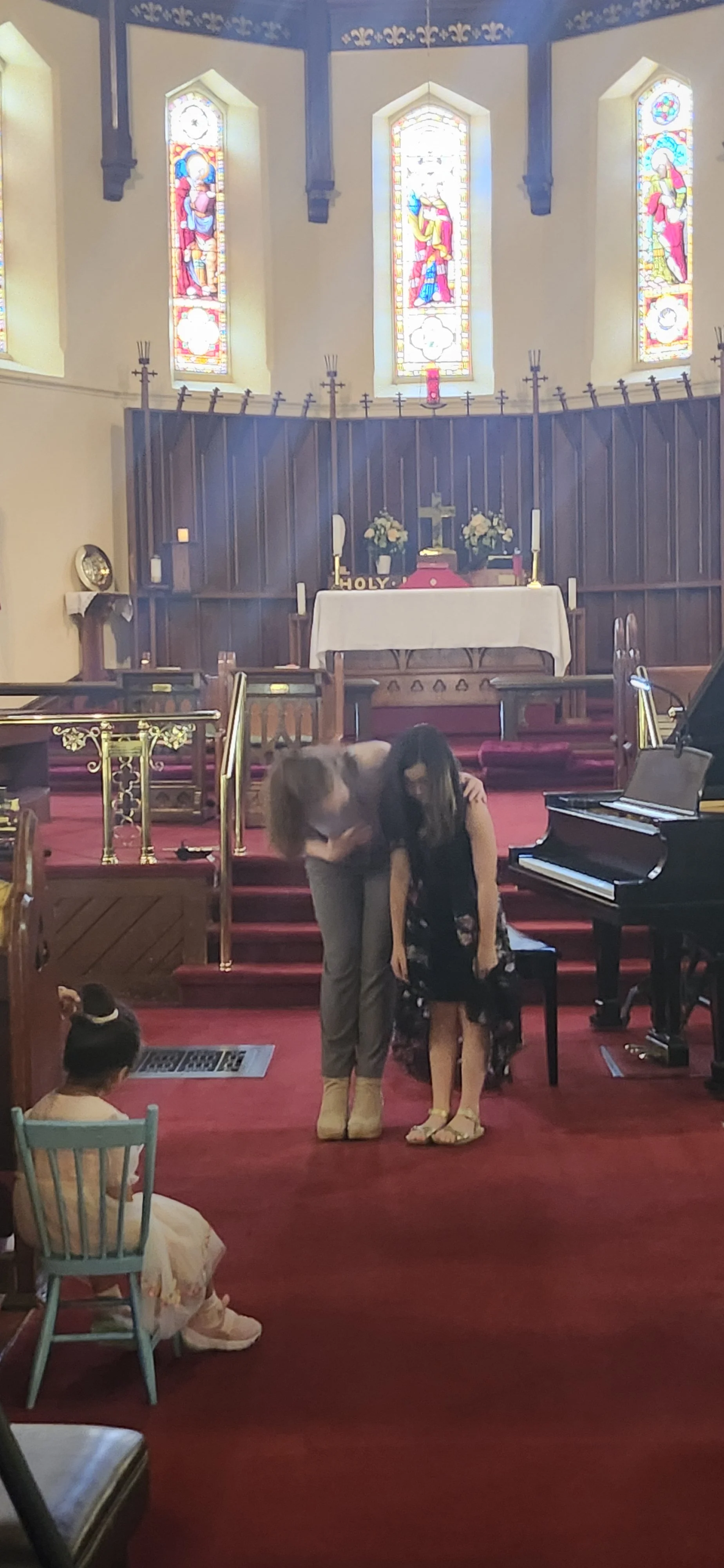 Two women bowing to each other inside a church with stained glass windows, an altar with candles, and a piano nearby. A child is sitting on a chair in the foreground.
