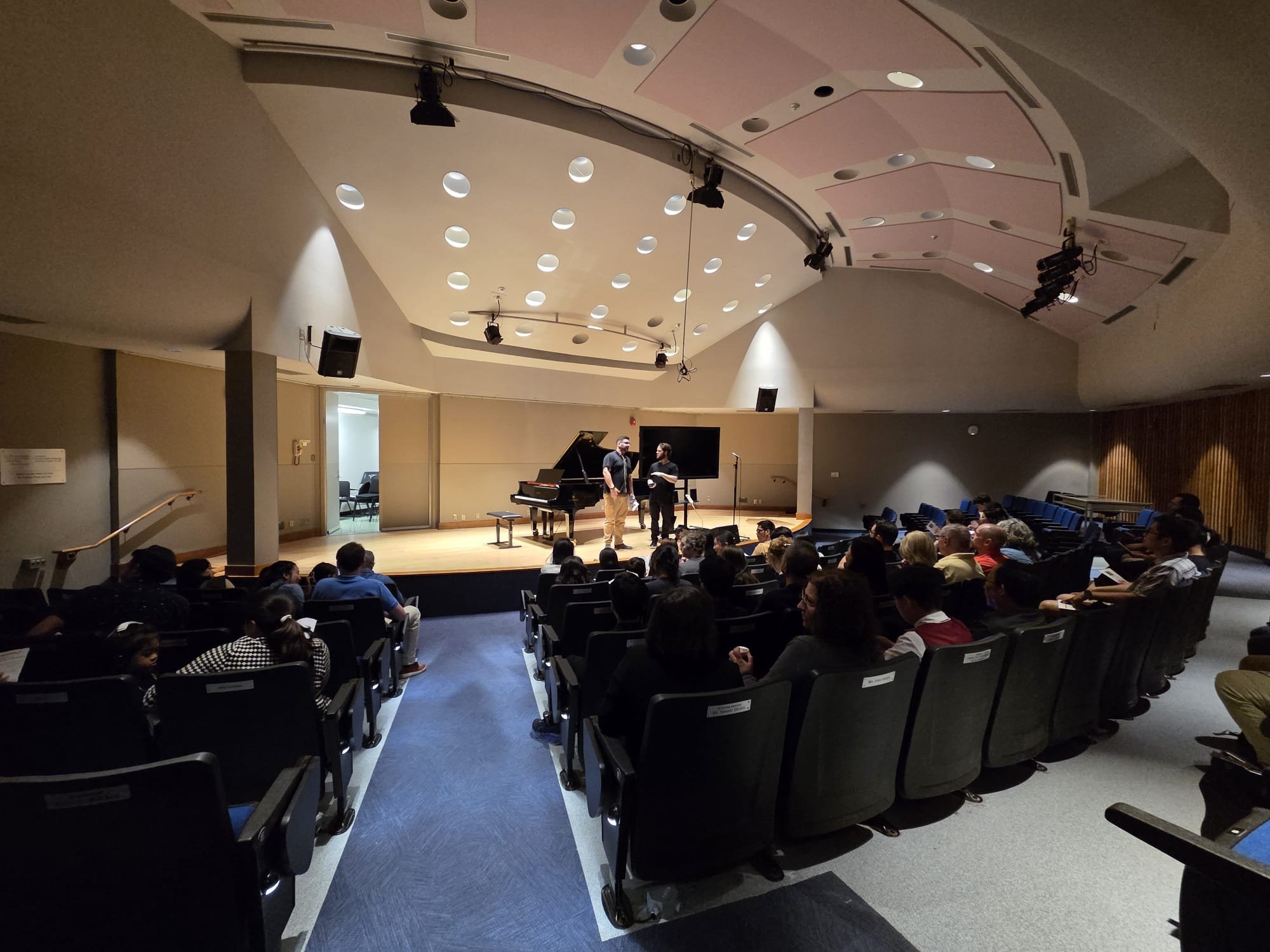 A concert hall or auditorium with a stage, where two people are standing and talking. There is a grand piano on the stage, and an audience seated in rows of chairs. The ceiling has circular lights and mounted speakers.